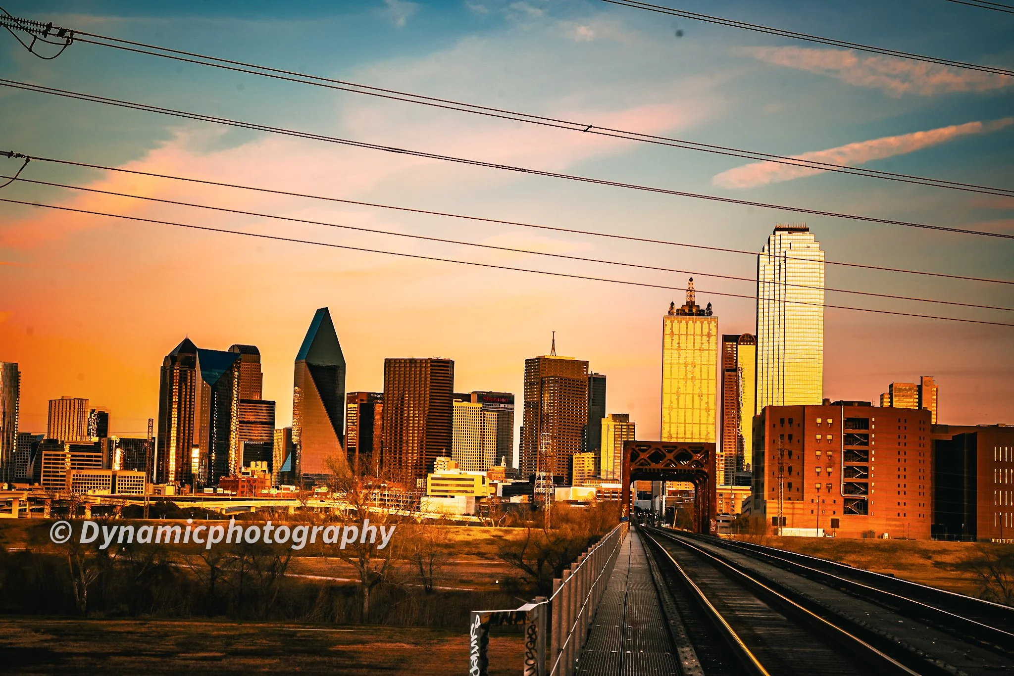 Sunset view of the Dallas city skyline with skyscrapers, train tracks, and power lines in the foreground.