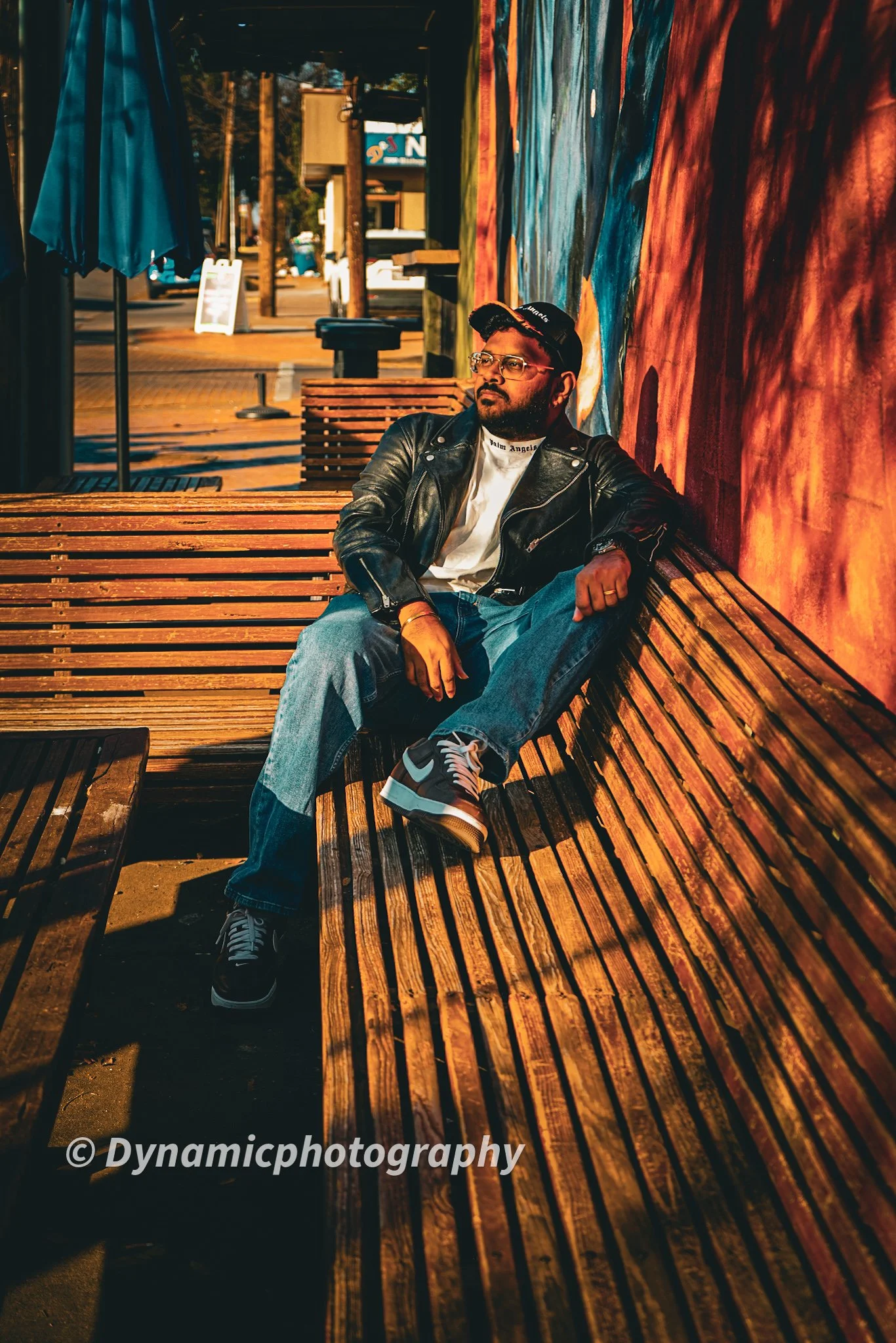 A man sitting on a wooden bench outside a colorful building, wearing a leather jacket, jeans, sneakers, sunglasses, and a baseball cap, with a mural wall behind him.