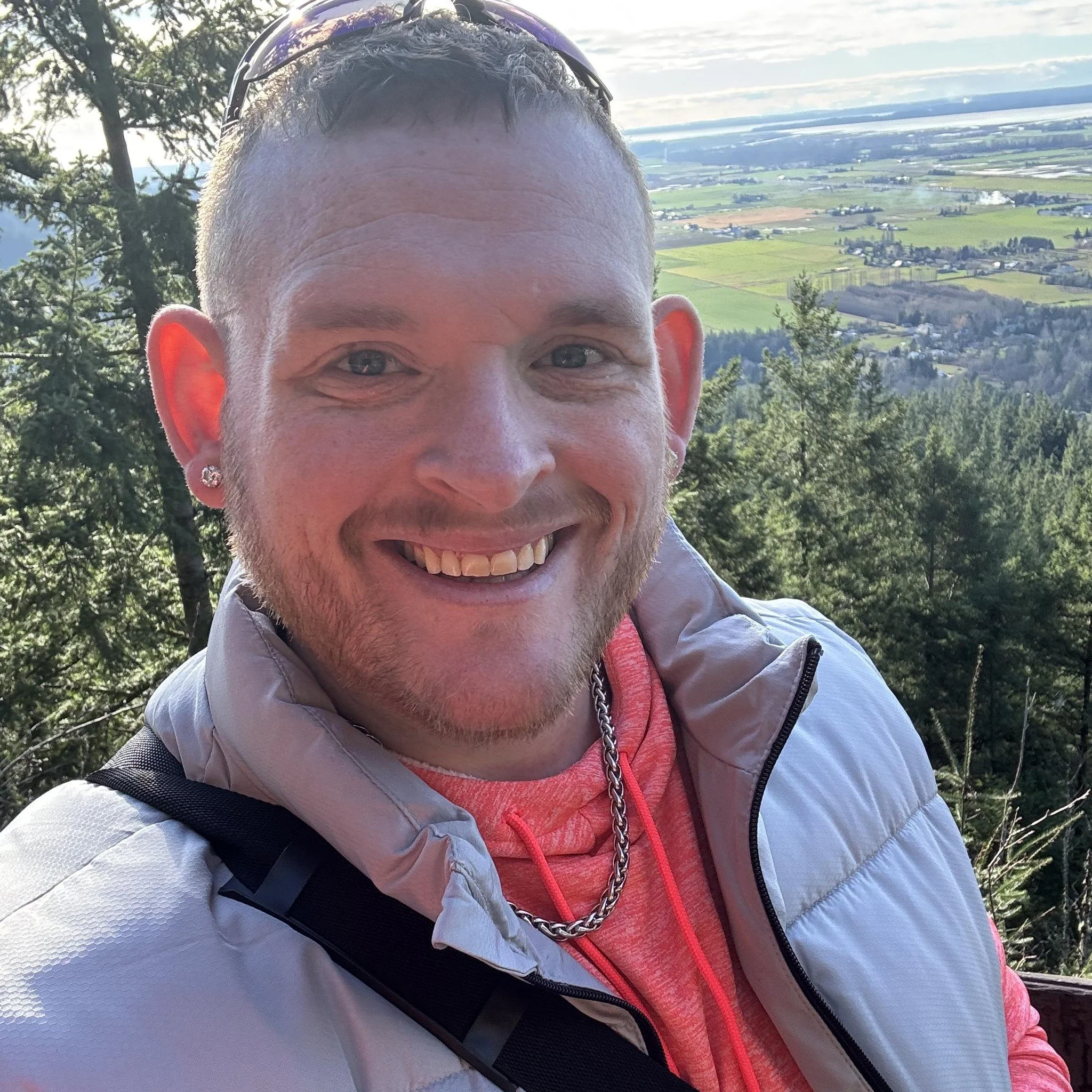A smiling man with short hair and a beard taking a selfie outdoors on a mountain trail, with a scenic view of trees and a valley in the background.