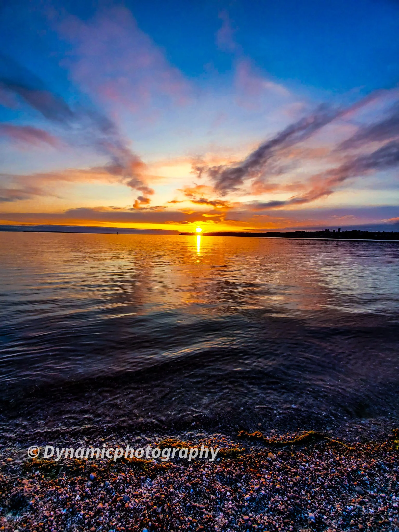 Sunset over water with a colorful sky and clouds, viewed from a pebbly shoreline.