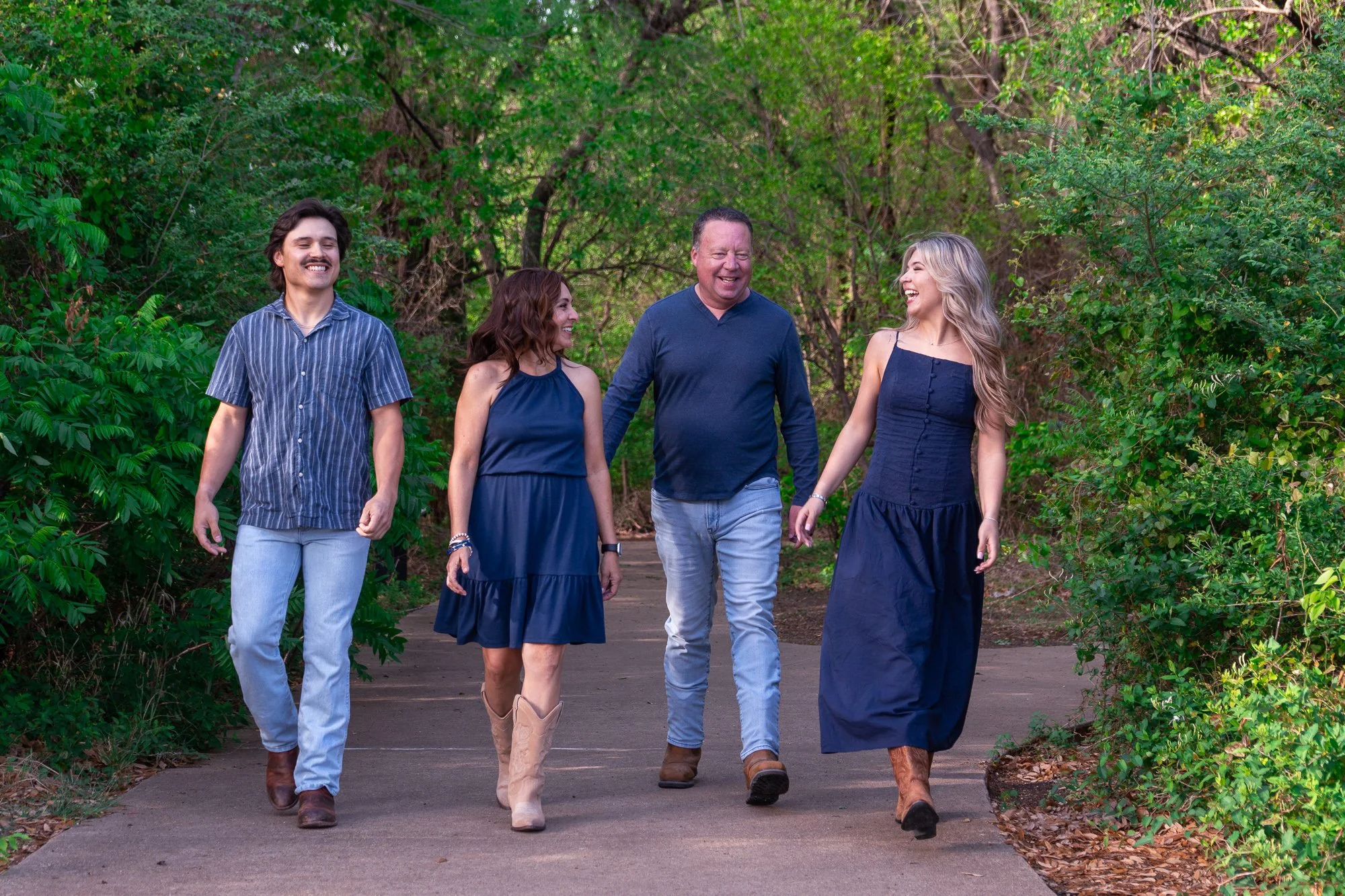 A happy family of four walking together on a wooded path, wearing coordinated navy blue outfits and cowboy boots, laughing and enjoying a candid moment during an outdoor lifestyle photography session.
