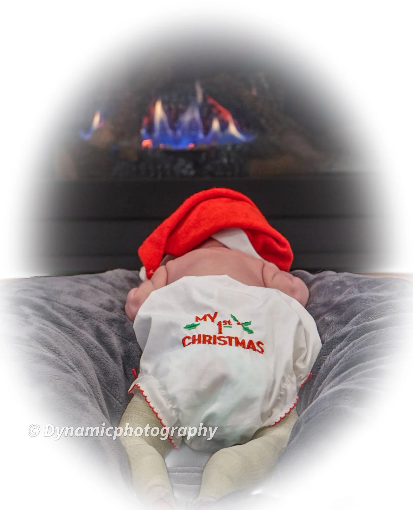 Baby lying face down in front of a fireplace wearing a Christmas-themed outfit, including a white cloth with red embroidery that says 'My 1st Christmas' and a red Santa hat.