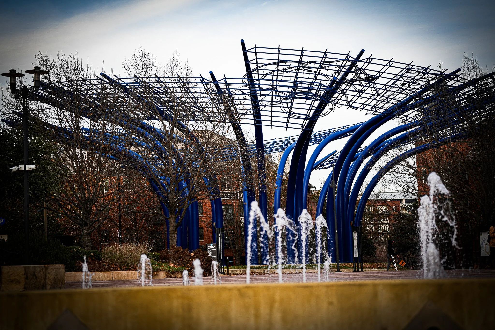 Public park with modern blue metal structure and water fountains in foreground, leafless trees, brick buildings, and a cloudy sky.