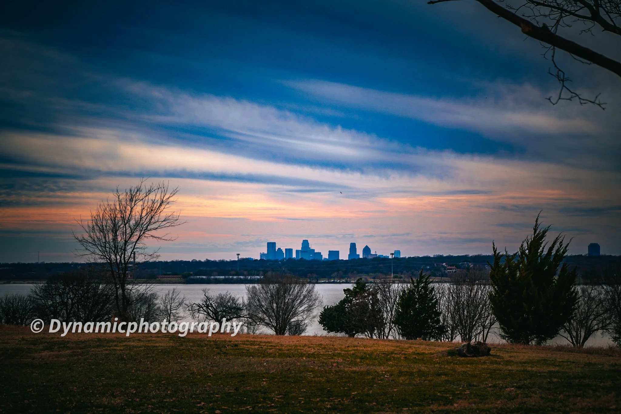 Sunset view of a city skyline across a body of water, with bare trees and grass in the foreground, and wispy clouds in the sky.