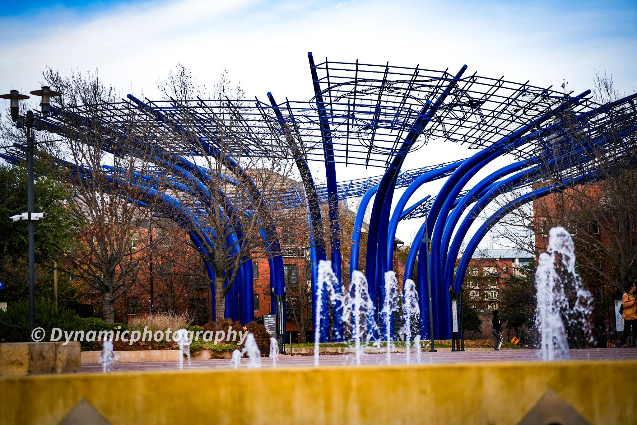 Public park with blue metal sculpture and water fountain, trees with no leaves, brick buildings in the background, cloudy sky.