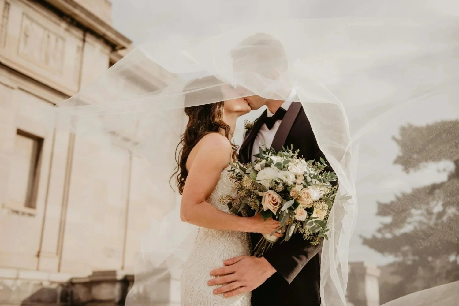 Bride and groom sharing a kiss under a wedding veil with a bouquet of flowers, outdoors near a white building.