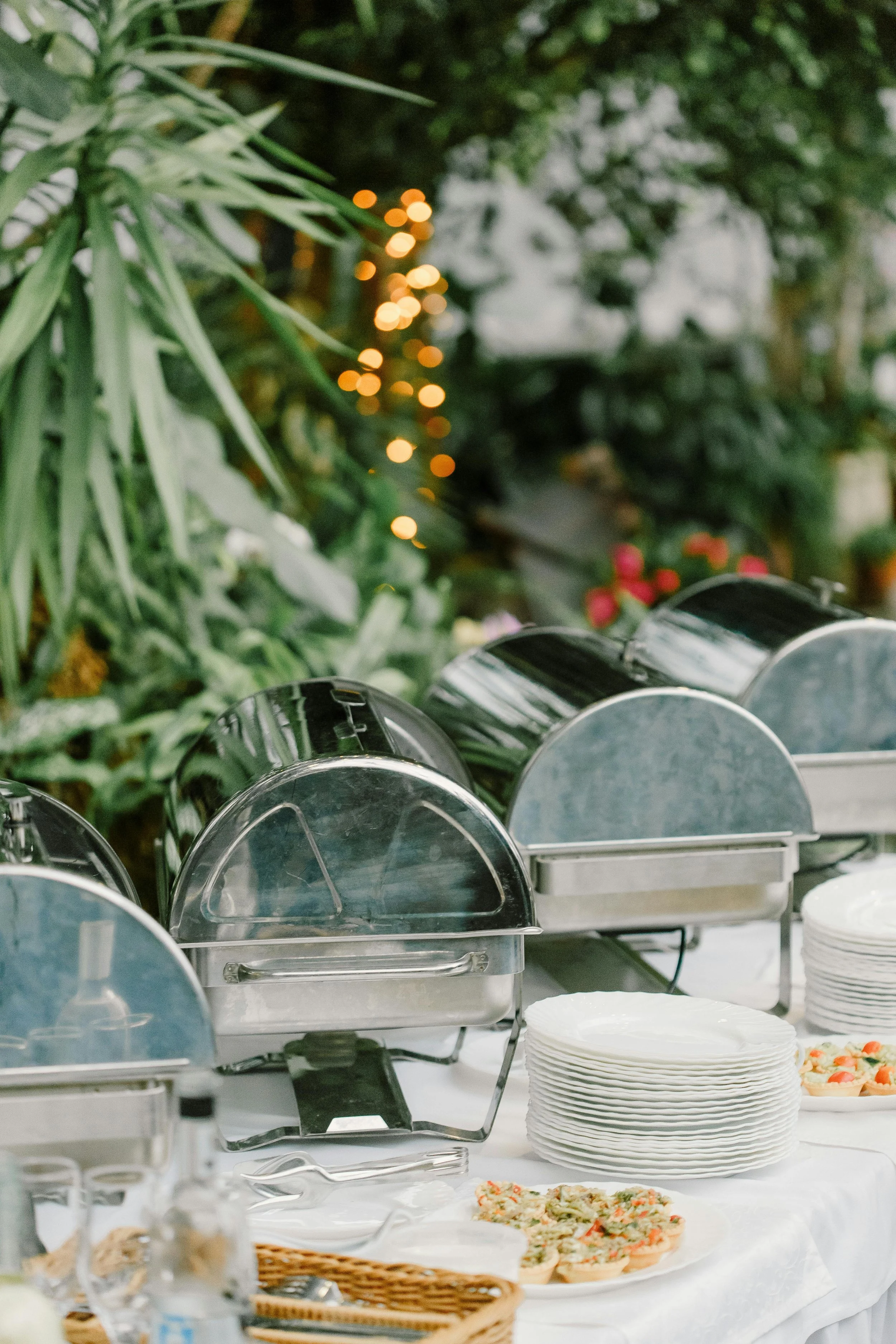 Buffet table with chafing dishes, a stack of white plates, and pizza slices, set outdoors with trees and string lights in the background.
