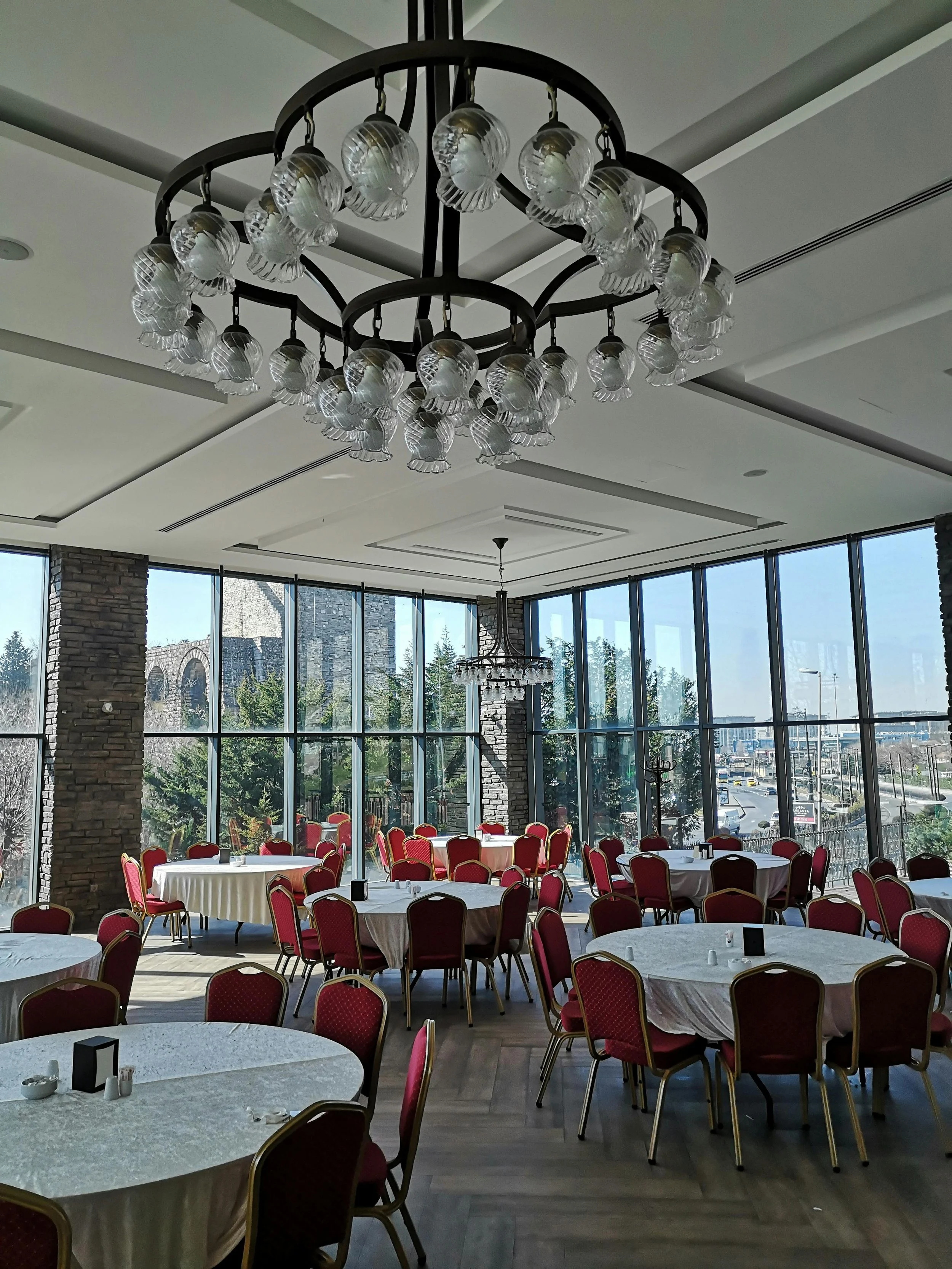 Elegant dining room with round tables covered in white tablecloths, red upholstered chairs, large glass windows, stone columns, and chandeliers.