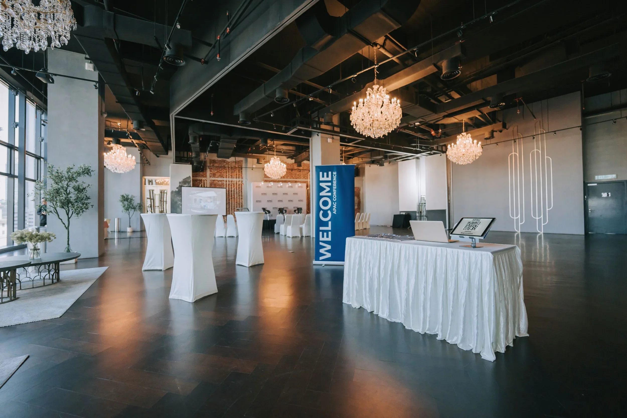 Modern event conference room with chandeliers, high ceilings, large windows, and a welcome banner, set up with standing tables and a registration table covered in white tablecloths.