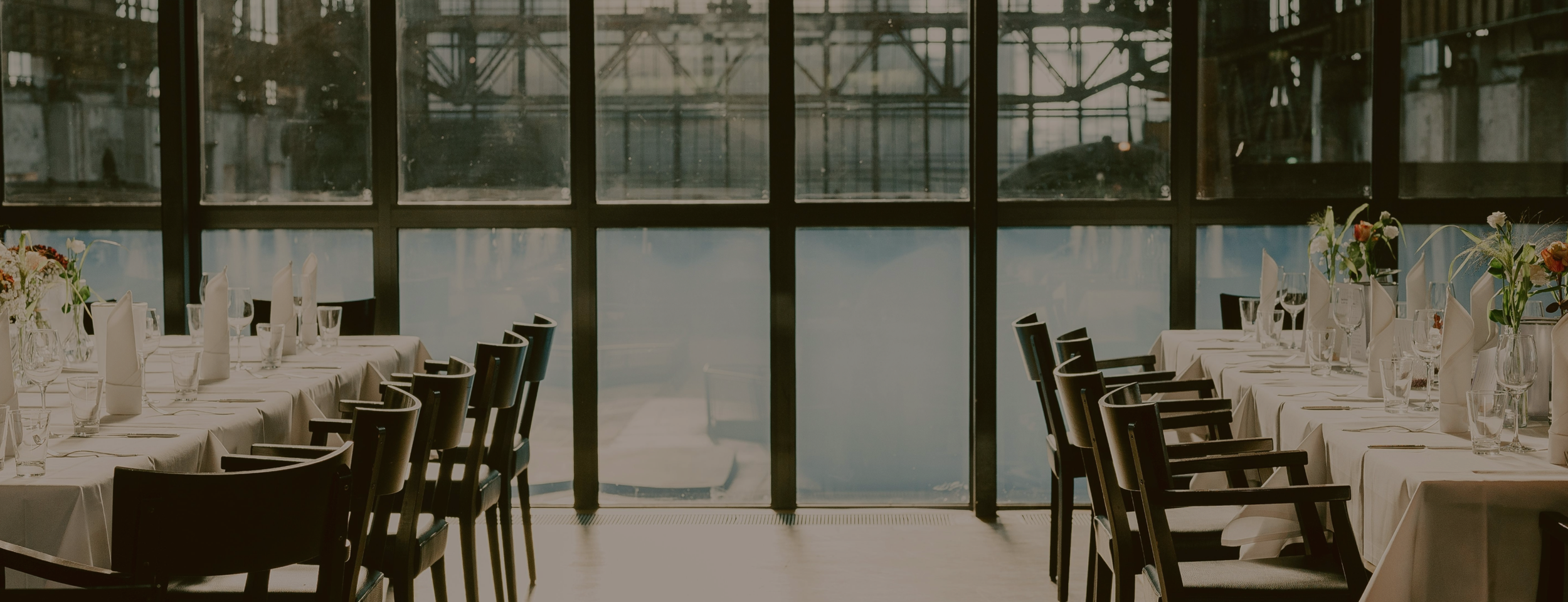 Empty restaurant dining area with white tablecloths, napkins, glasses, and vases with flowers, overlooking a large window with an exterior view of an airport runway or hangar.