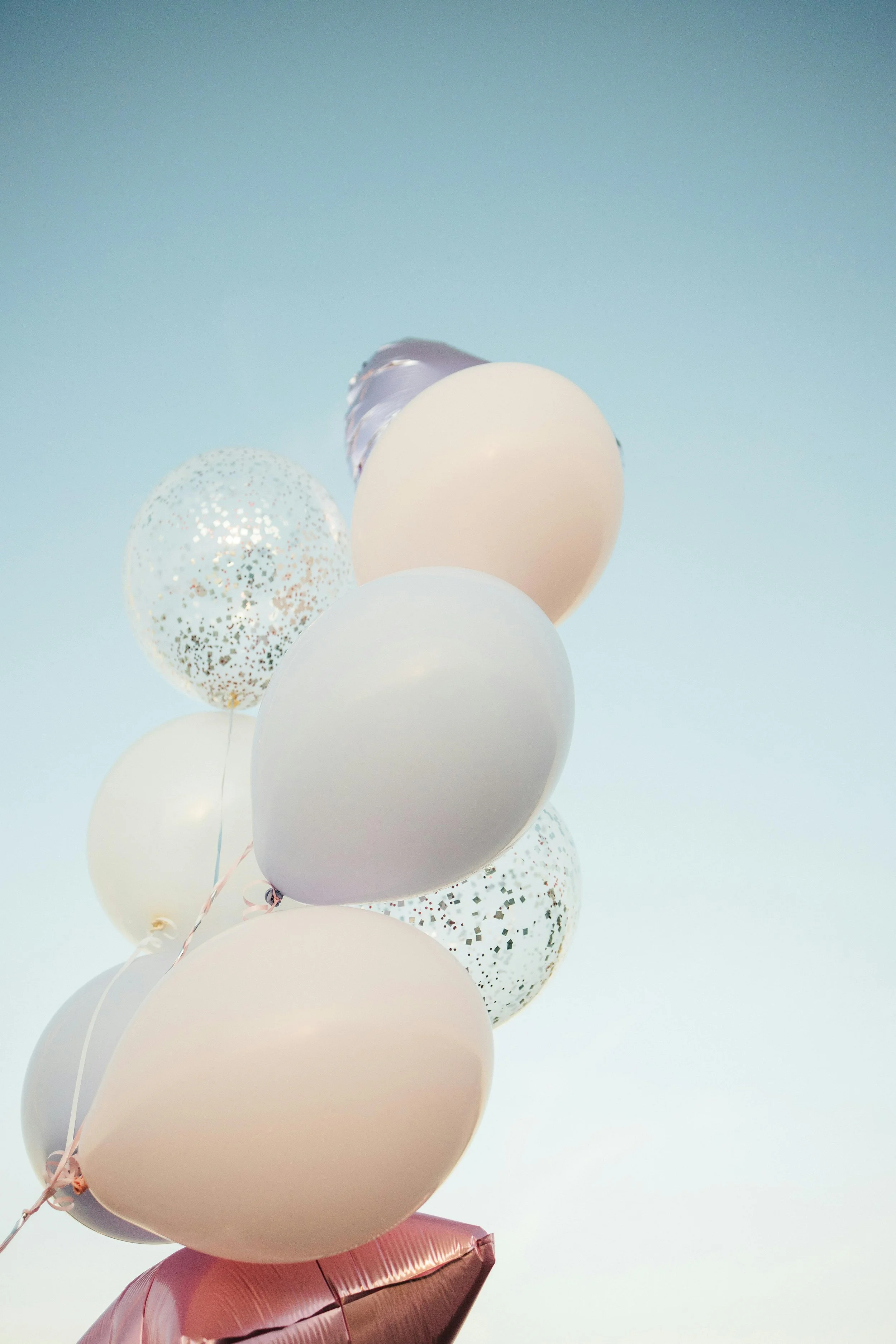 A cluster of balloons, including white, pastel pink, with glitter and metallic finishes, against a pale blue sky.