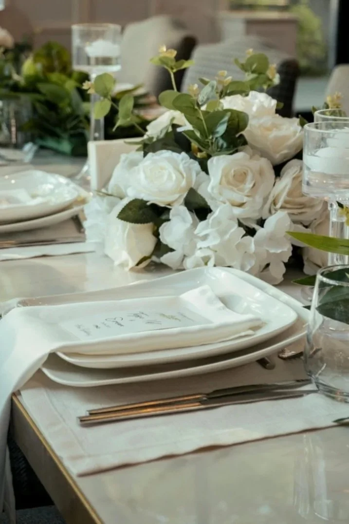 Elegant table setting with white plates, silverware, glassware, and a floral centerpiece of white roses and green leaves.