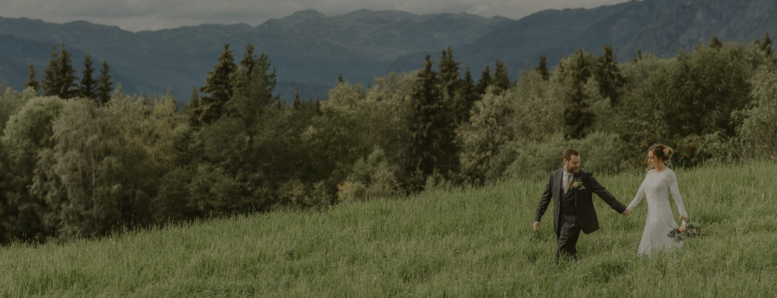 A bride and groom holding hands walking in a grassy field with trees and mountains in the background.