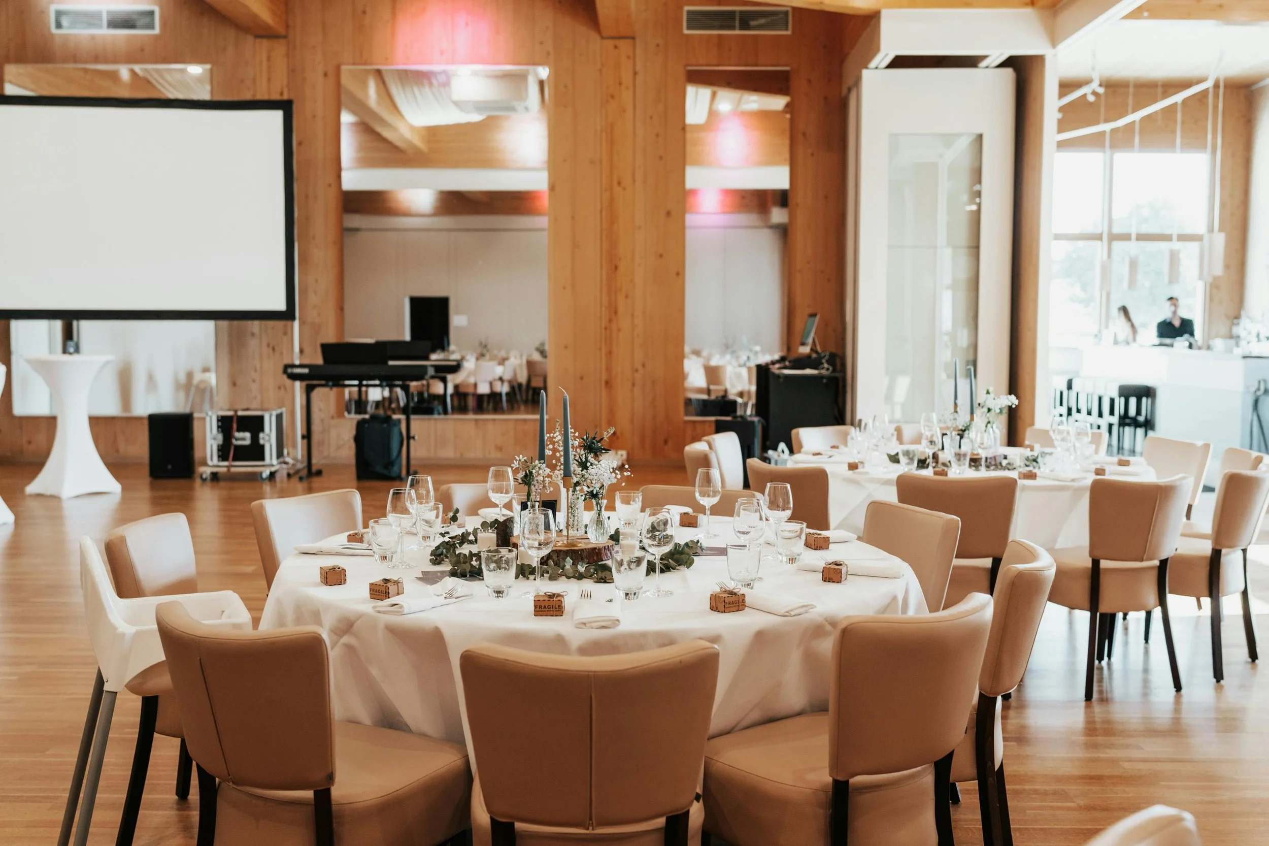 Event space set up for a wedding reception or formal gathering with decorated round tables, wine glasses, candles, and floral centerpieces in a wooden interior.