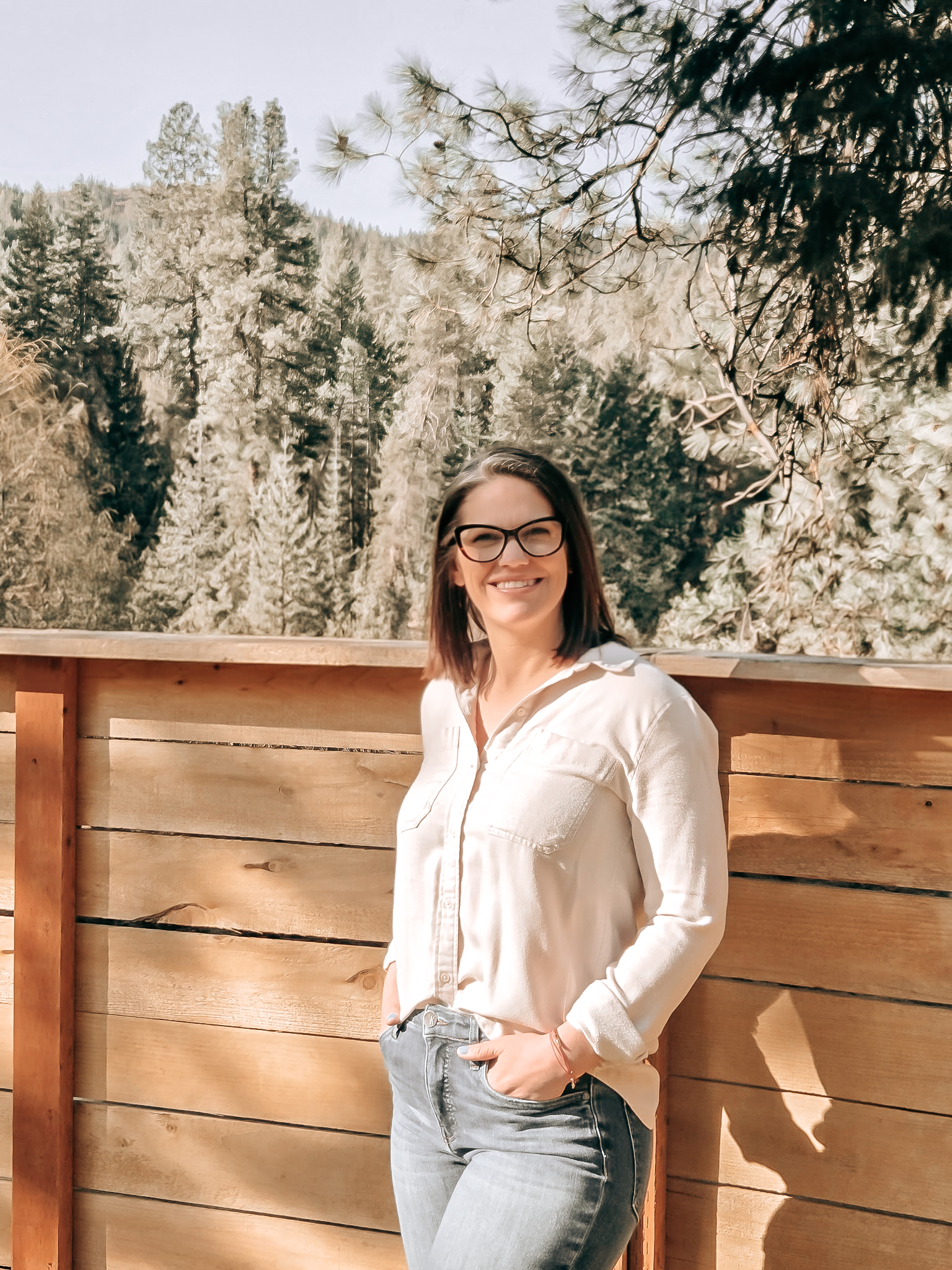 A woman with glasses and wearing a white shirt and gray jeans, smiling while standing outdoors near a wooden fence with a forest background.