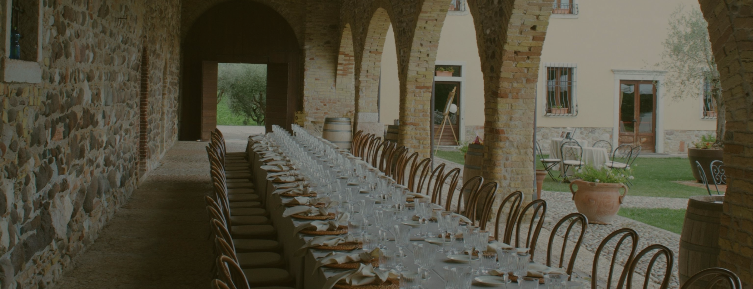 Long dining table set with white napkins, glassware, and dinnerware, under a stone and brick archway, with outdoor garden visible in the background.