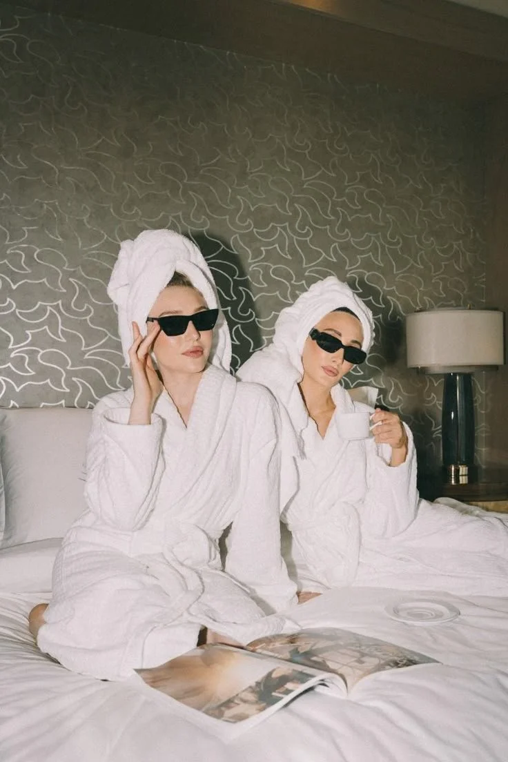 Two women in bathrobes and towels wrapped around their heads, wearing sunglasses, sitting on a bed, one holding a cup and the other touching her sunglasses, with a magazine and a CD on the bed.