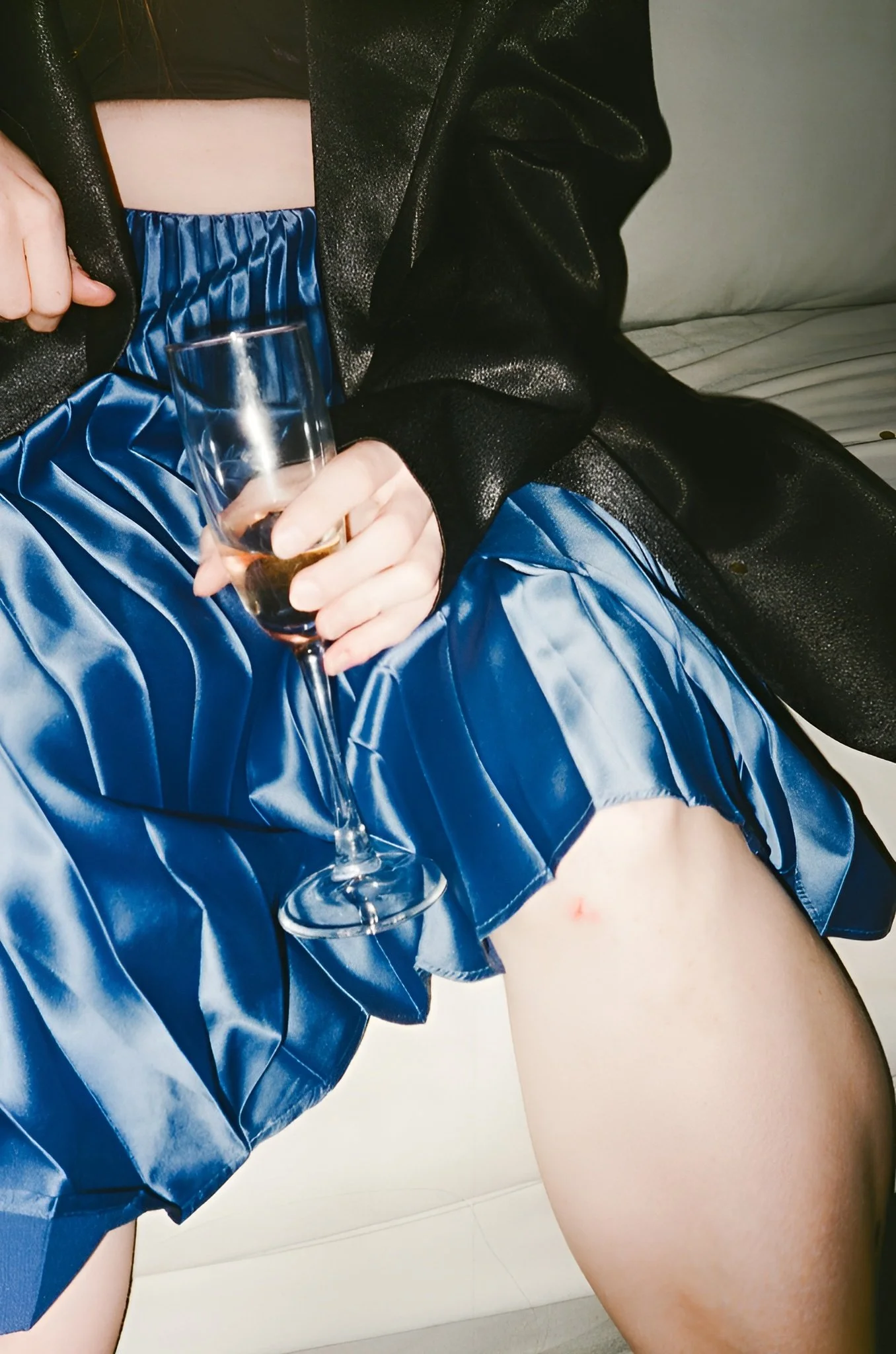 Person wearing a black blazer and blue satin skirt, holding a glass of wine, sitting on a white couch.