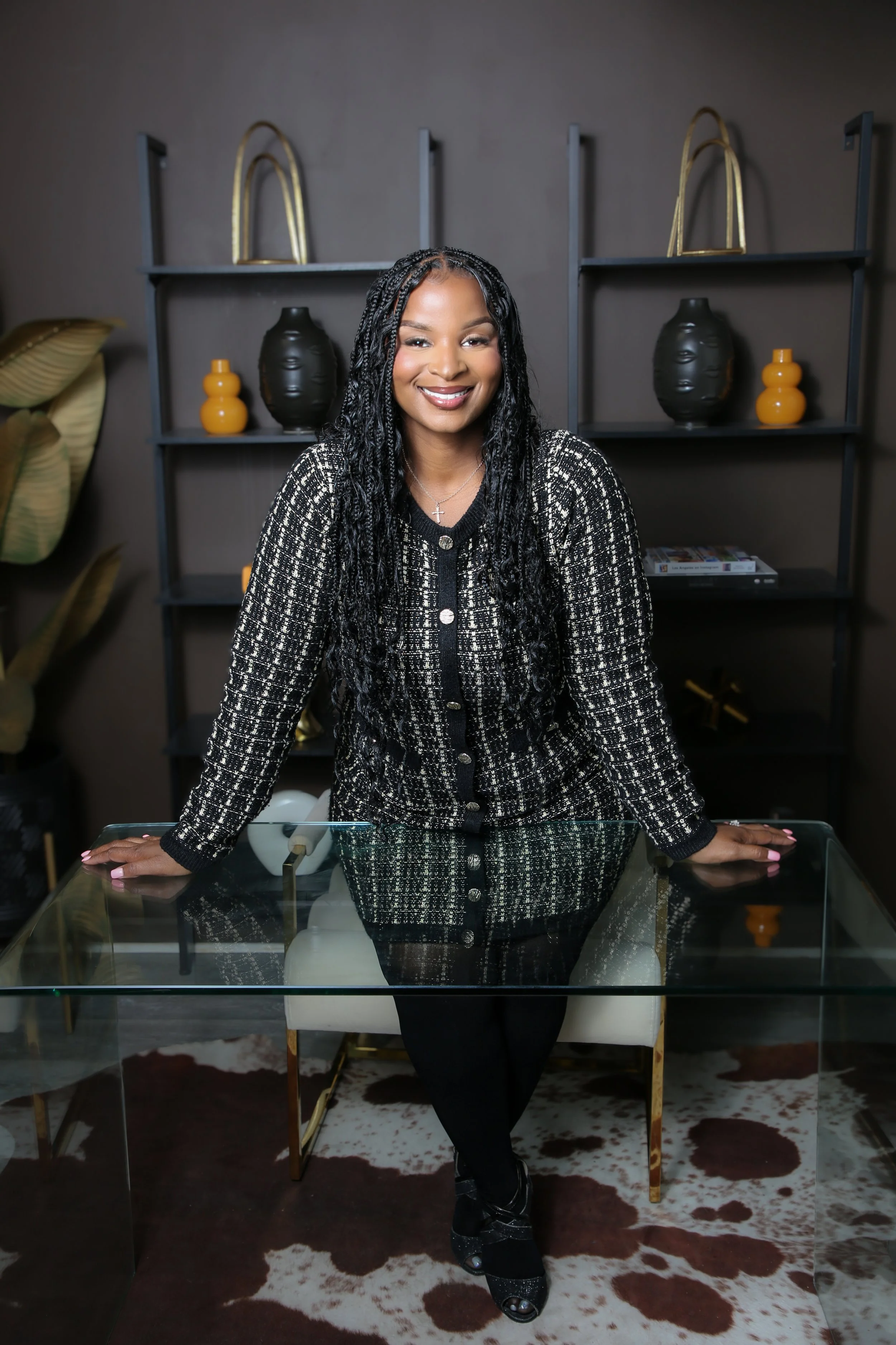 A woman with long, twisted black hair, wearing a black and white checkered jacket, leaning on a glass table in a modern room with decorative vases and wall shelves.