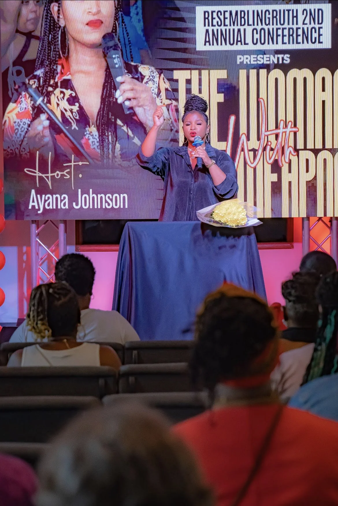 A woman speaking on stage during the Rebuilding Ruth 2nd Annual Conference, with a large screen behind her displaying her name as Ayana Johnson and the event title. She is holding a microphone and standing behind a table with a bouquet of flowers.
