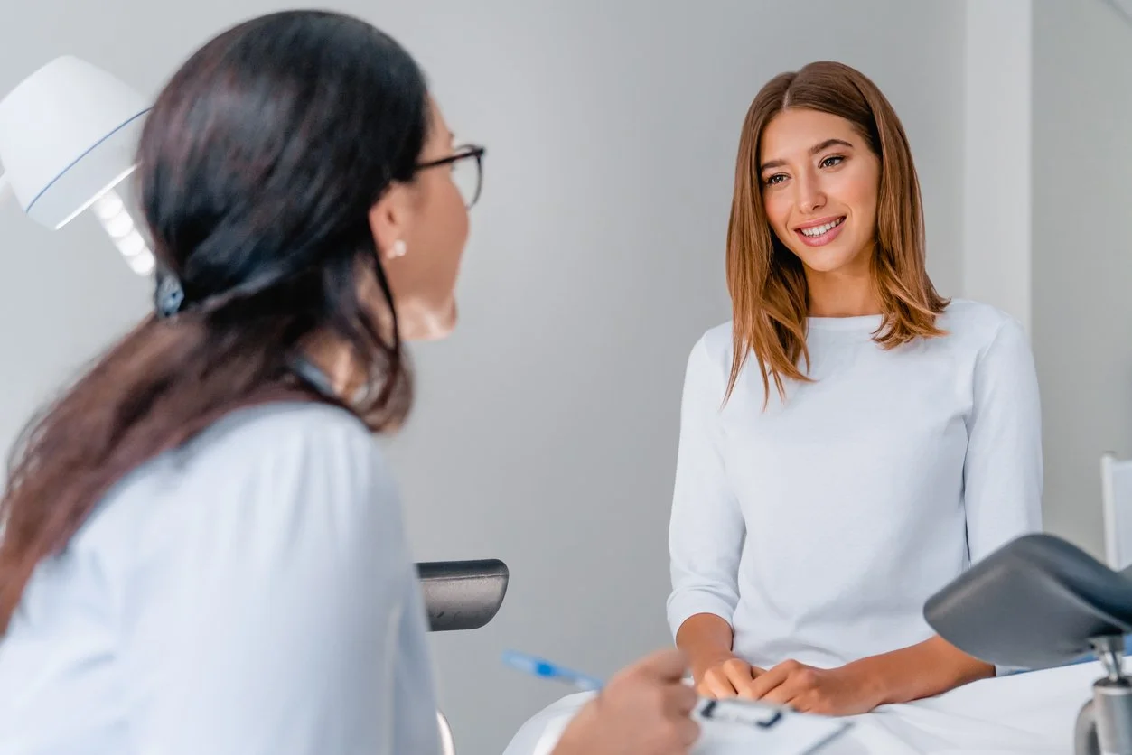 Young woman in white shirt sitting in a medical examination room, talking with a female healthcare professional who is writing notes.