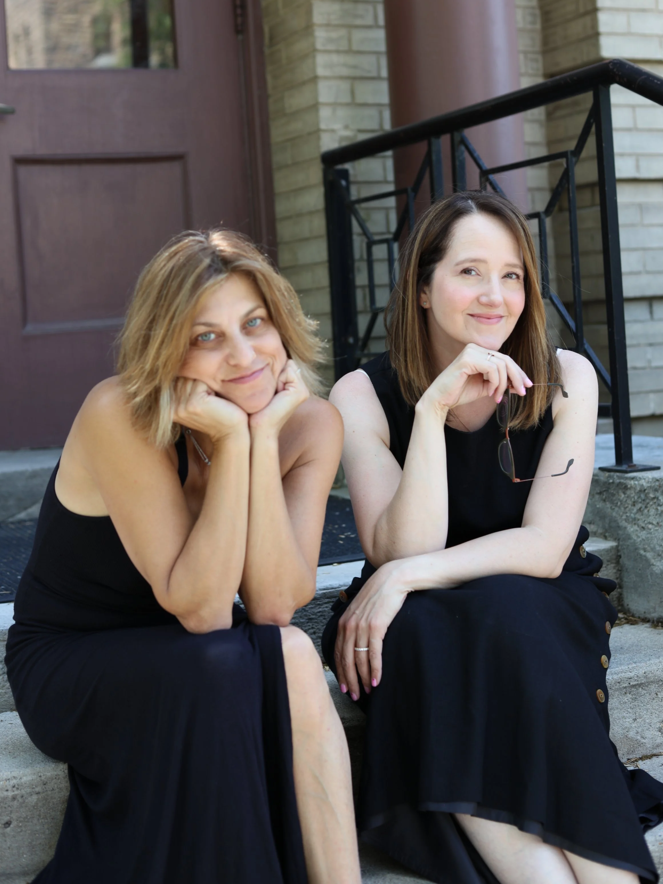 Two women sitting on stairs outside a building, smiling at the camera. One has blonde hair and the other has brown hair, both wearing black dresses.