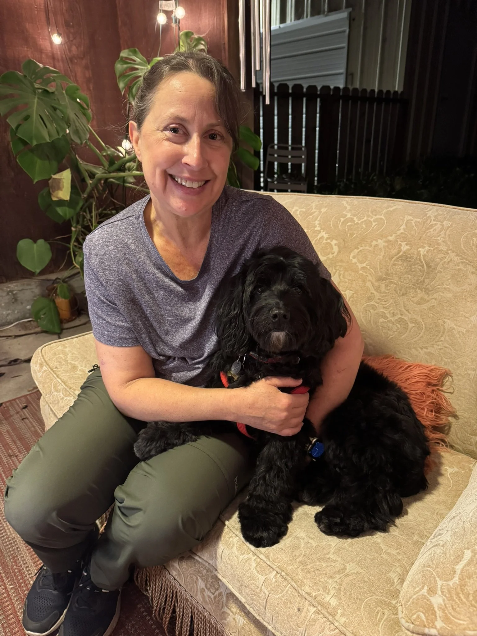 A woman with short brown hair smiling while holding a black puppy on her lap, sitting on a beige patterned couch in an outdoor setting at night.