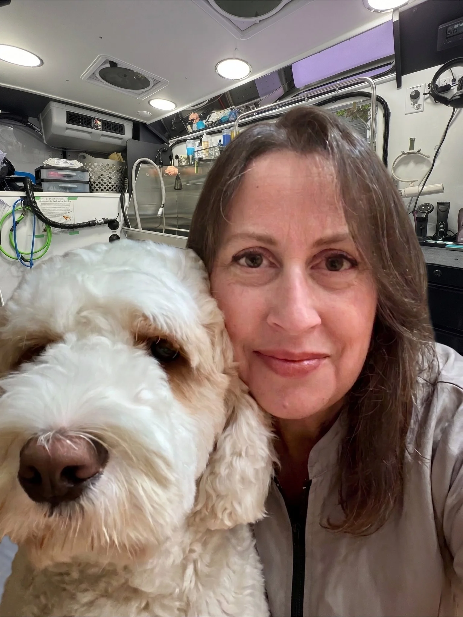 A woman with brown hair and a smile taking a selfie with a fluffy white dog inside a medical or laboratory vehicle or setting.