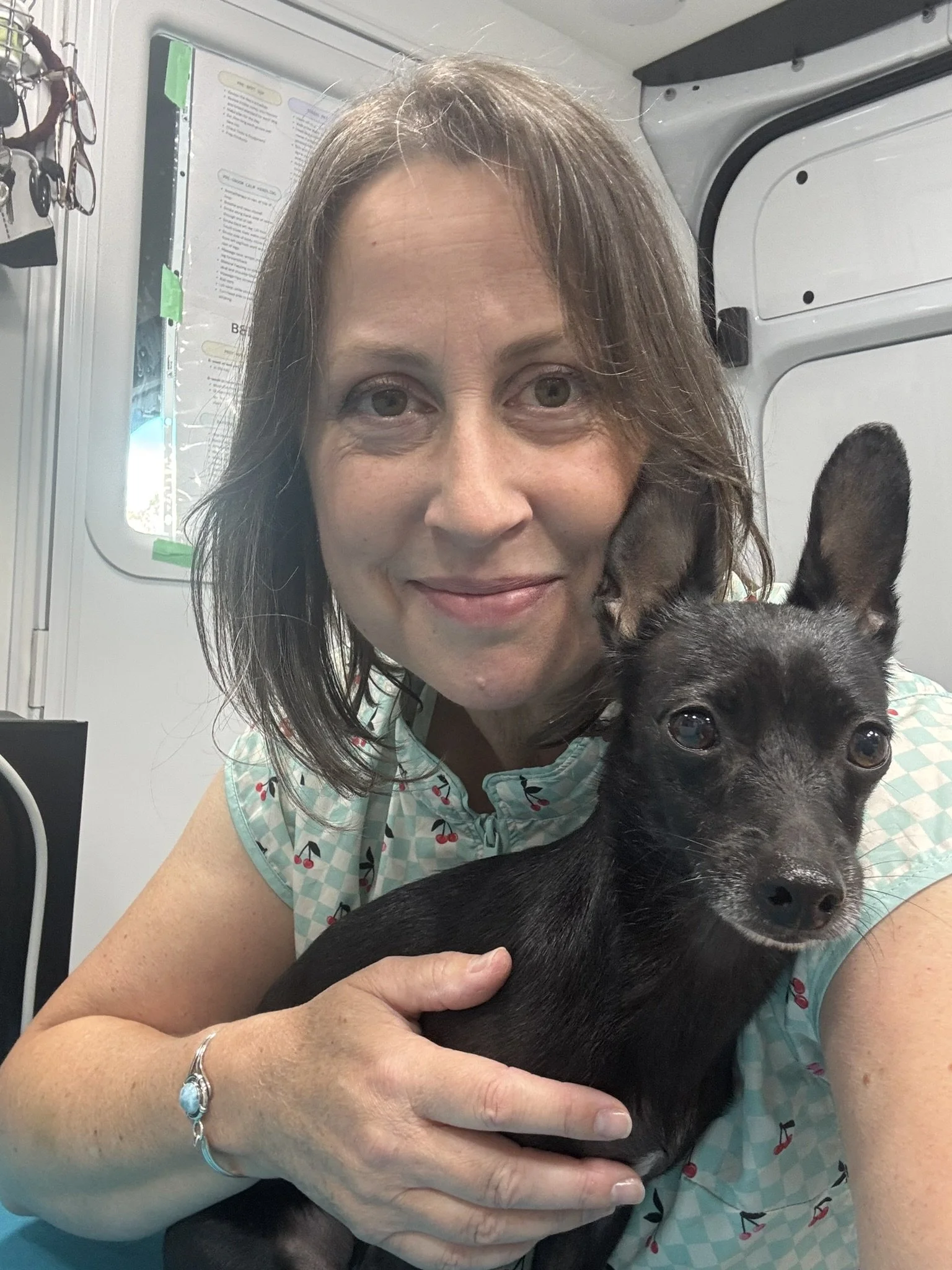 A woman in a light blue dress with a cherry pattern is holding a black puppy with pointy ears. She is smiling at the camera, and they are indoors, possibly in a veterinary clinic or animal shelter.