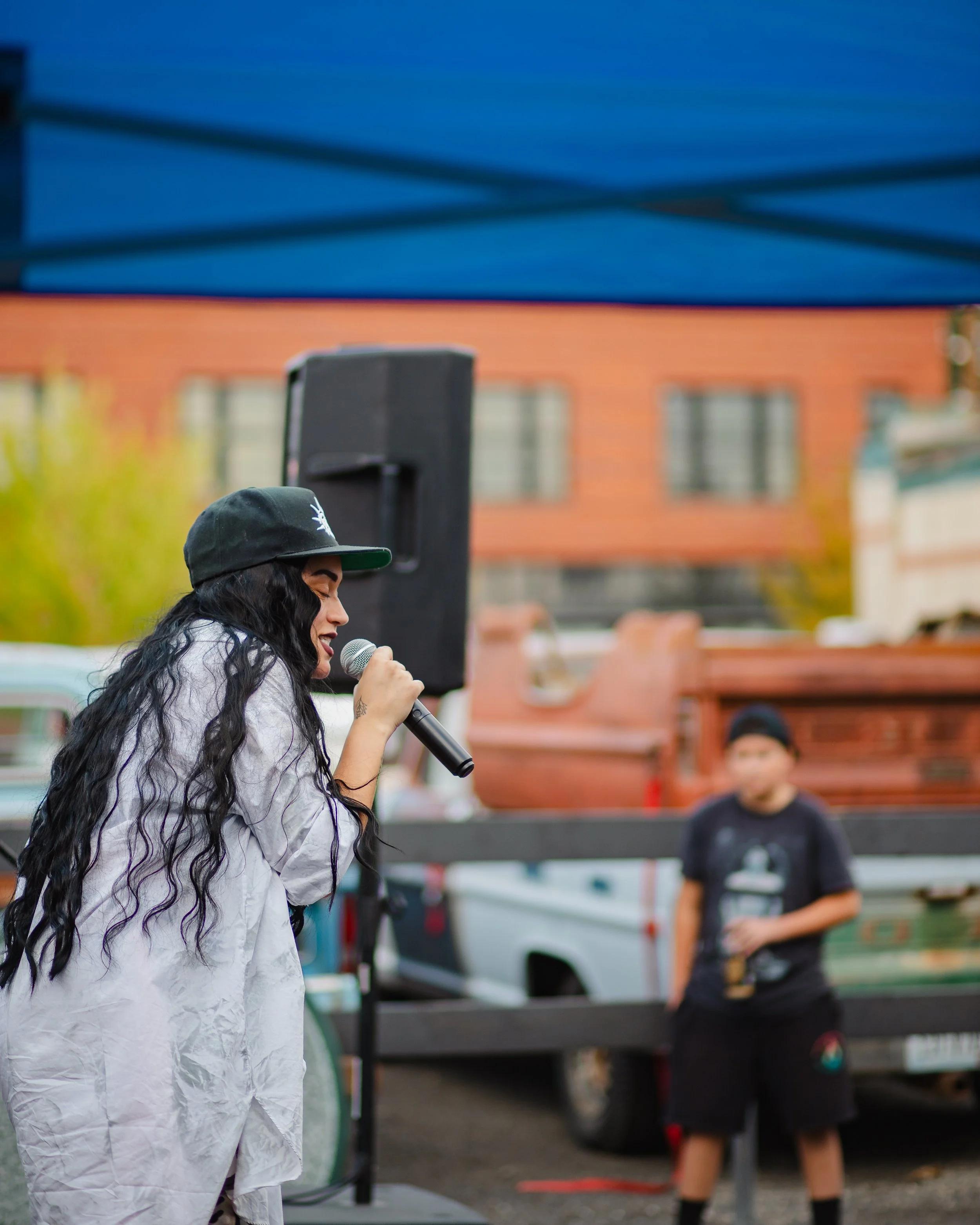HNY with long black hair wearing a black cap and a white oversized shirt singing into a microphone outdoors at Bodega Block Party. A man stands in the background holding a cup, with trucks and buildings behind them.