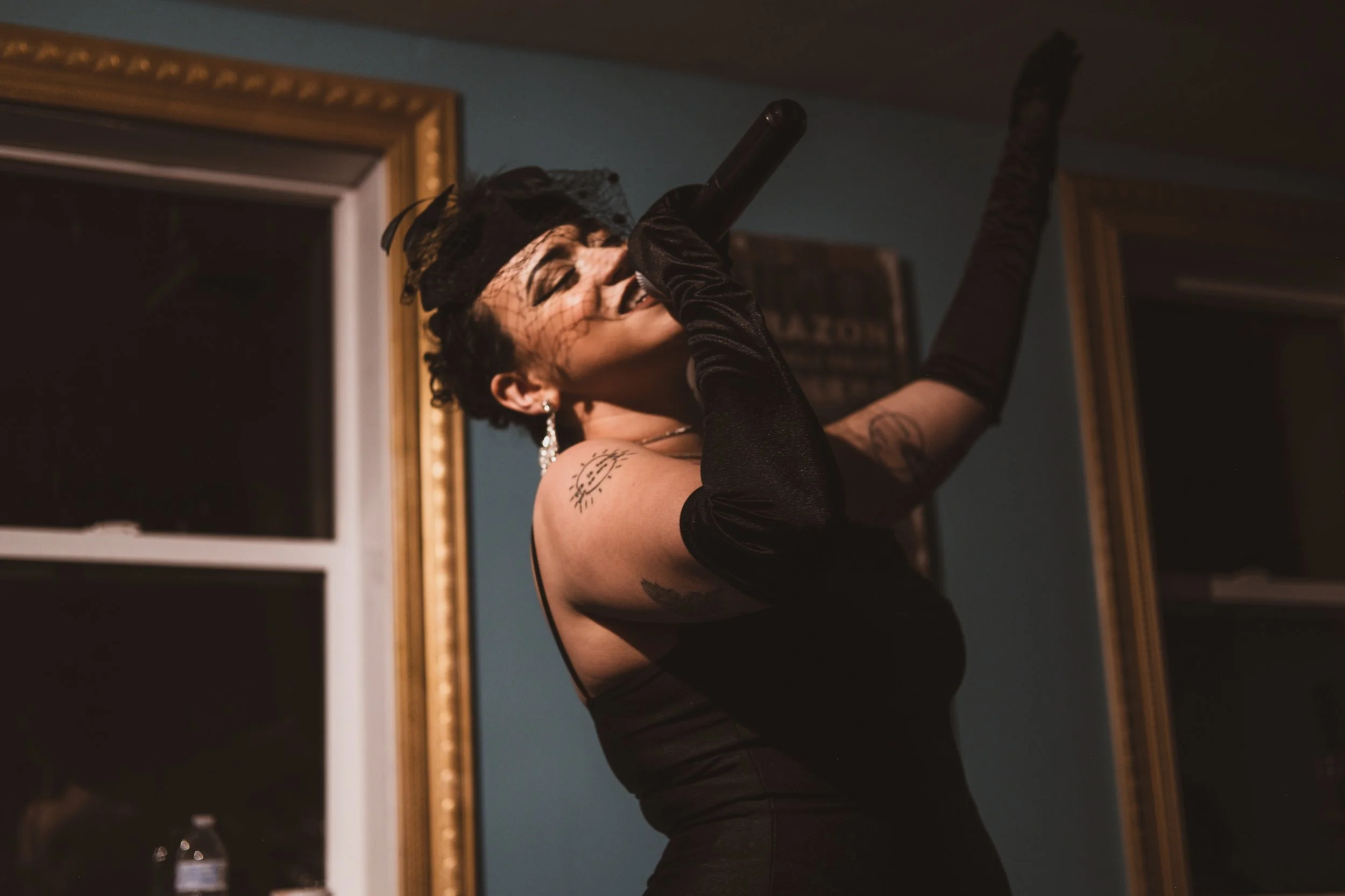 HNY in a black dress and long black gloves singing into a microphone, with a black veil on her head, in a room with mirrors and blue walls.