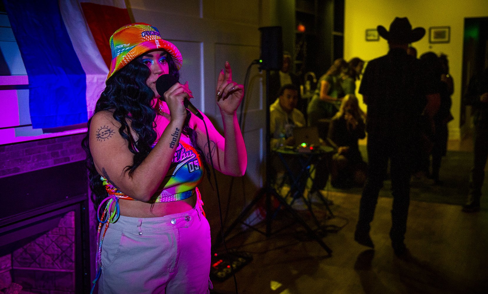 A woman with long black curly hair, wearing a rainbow bucket hat, colorful top, and white pants, singing into a microphone at a lively party or event. There is a rainbow flag hanging behind her, and several people are in the background, some standing and some sitting, with dim lighting and colorful illumination.