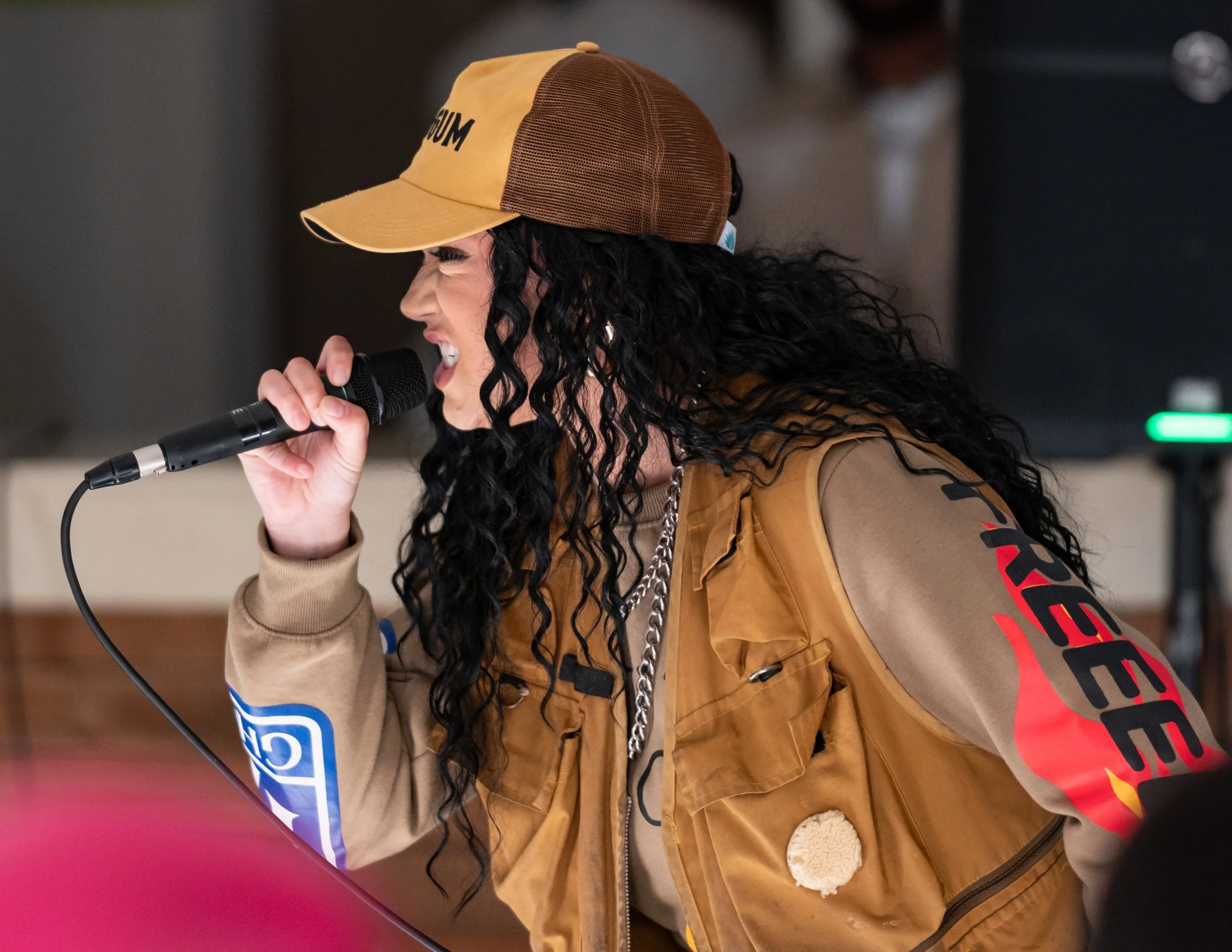 HNY with curly black hair wearing a tan and brown baseball cap, a brown vest with patches, and a beige shirt, rapping into a microphone.