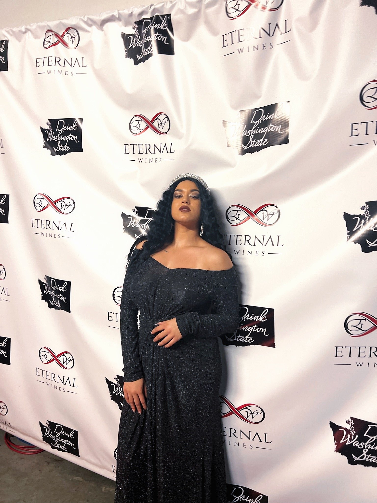 HNY with dark curly hair and dark lipstick wearing a long, sparkly black off-the-shoulder gown standing against a step-and-repeat backdrop. The backdrop has logos for Eternal Wines and Drink Washington State.