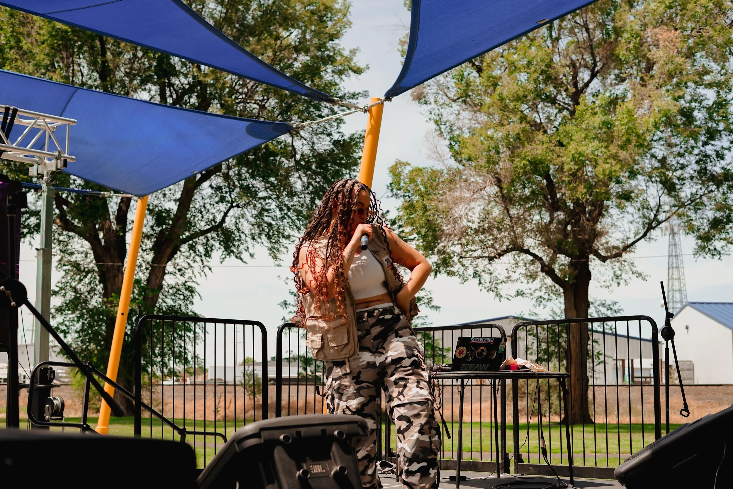 HNY with long, curly hair performing on an outdoor stage, holding a microphone. She is wearing camouflage pants and a beige vest with a white crop top. The stage is covered by large blue shade sails, with trees and buildings in the background.
