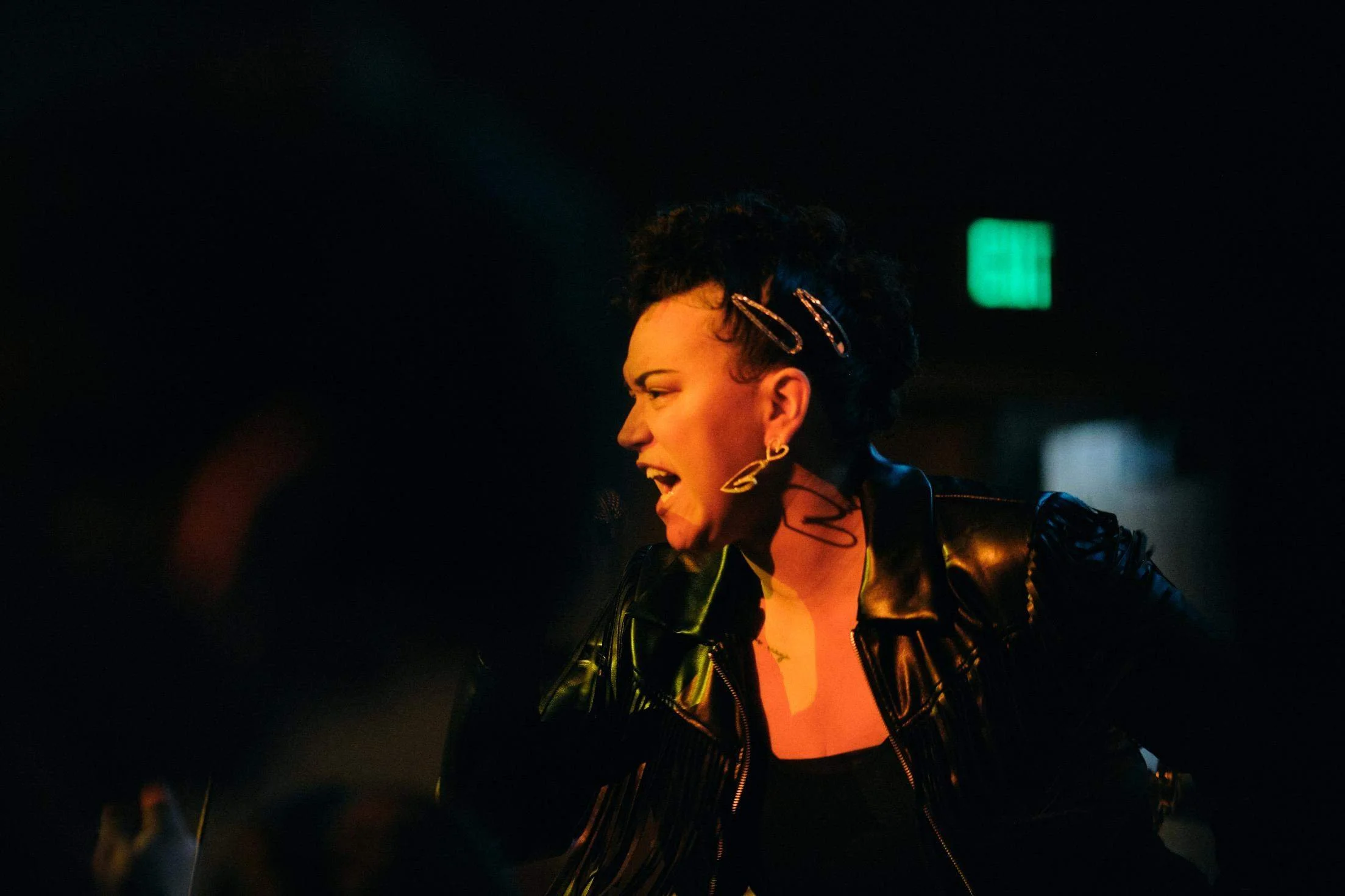 HNY performing passionately, wearing a black leather jacket, with short curly hair held back with hair clips, and large earrings. The background is dark with a green exit sign.