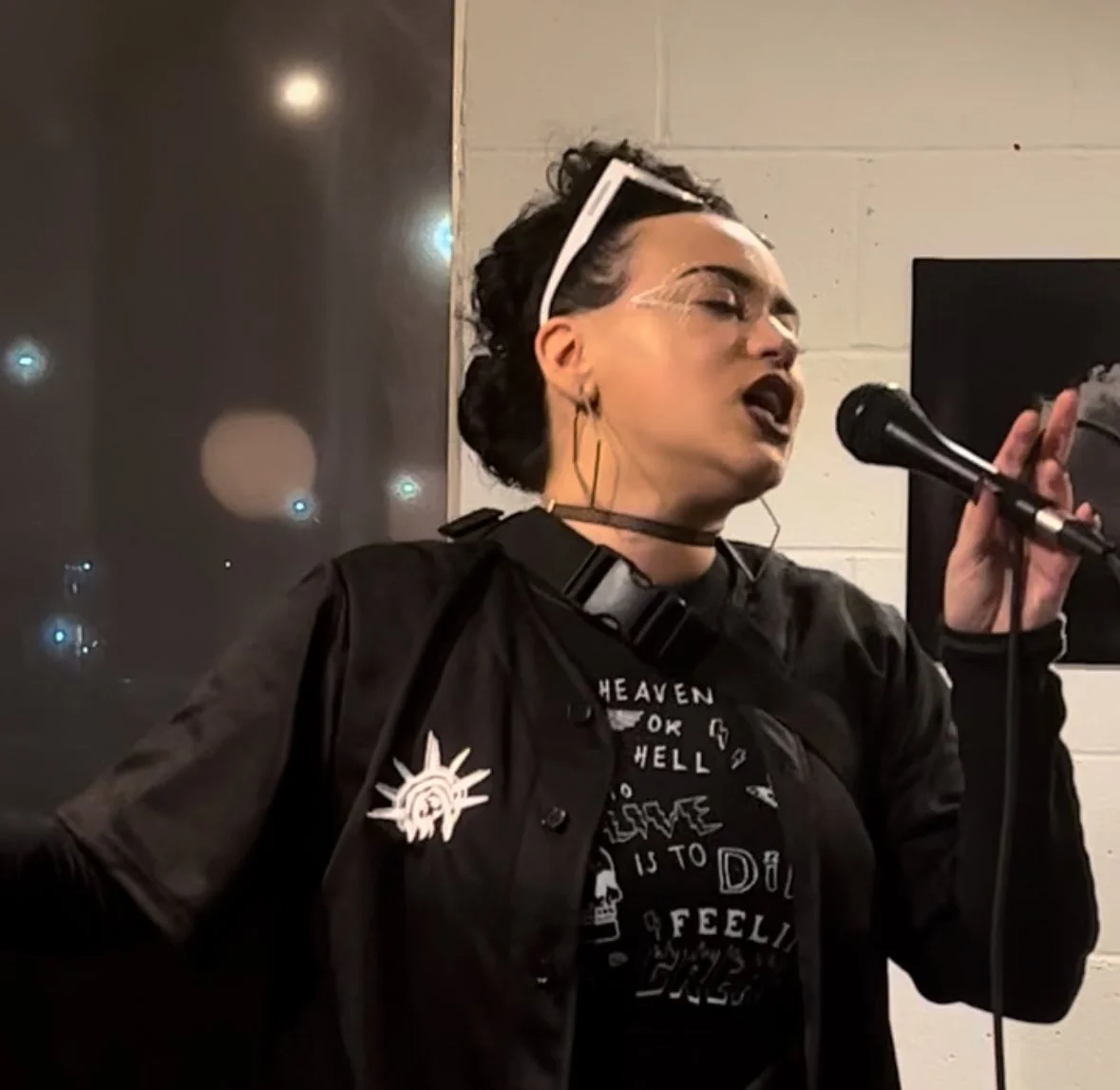 HNY singing into a microphone at FVC Gallery, wearing glasses on her forehead, a black jacket with pins, and a graphic t-shirt, with white brick walls in the background.