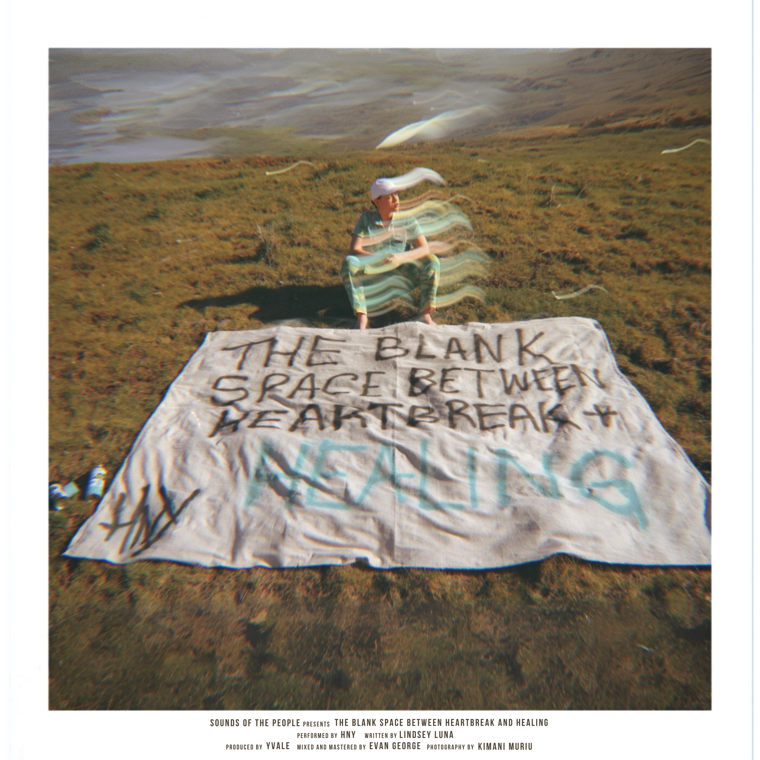A person sitting on a grassy shoreline behind a large, handmade sign that reads, 'The blank space between heartbreak + healing.' The sign appears to be made from fabric and is laid on the ground. The person is wearing colorful clothing and a white cap, with an ocean or lake in the background.