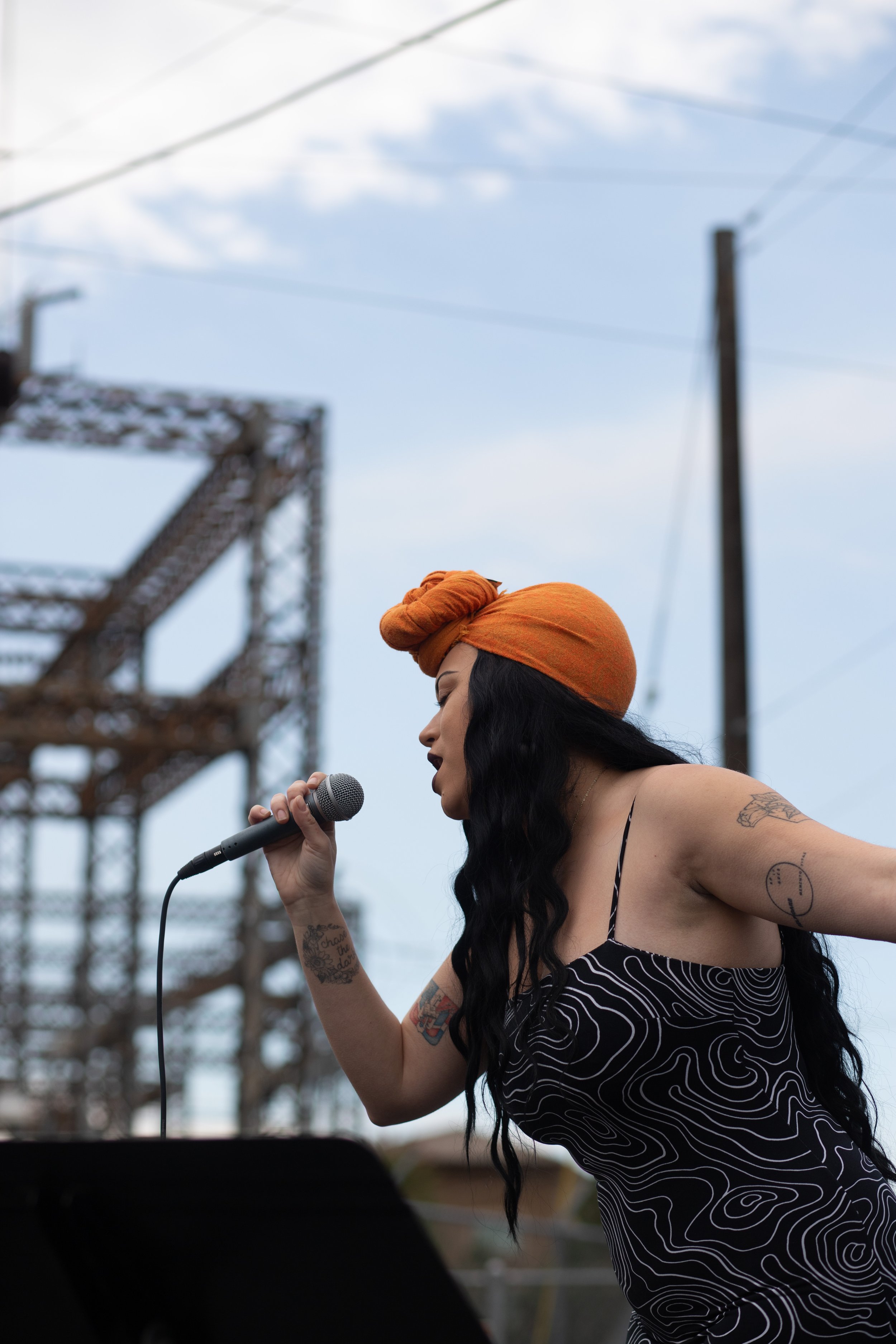 HNY with long black hair, tattoos on her arms, wearing a black and white patterned dress and an orange headwrap, singing into a microphone outdoors at Gesa Powerhouse Theatre under a partly cloudy sky.