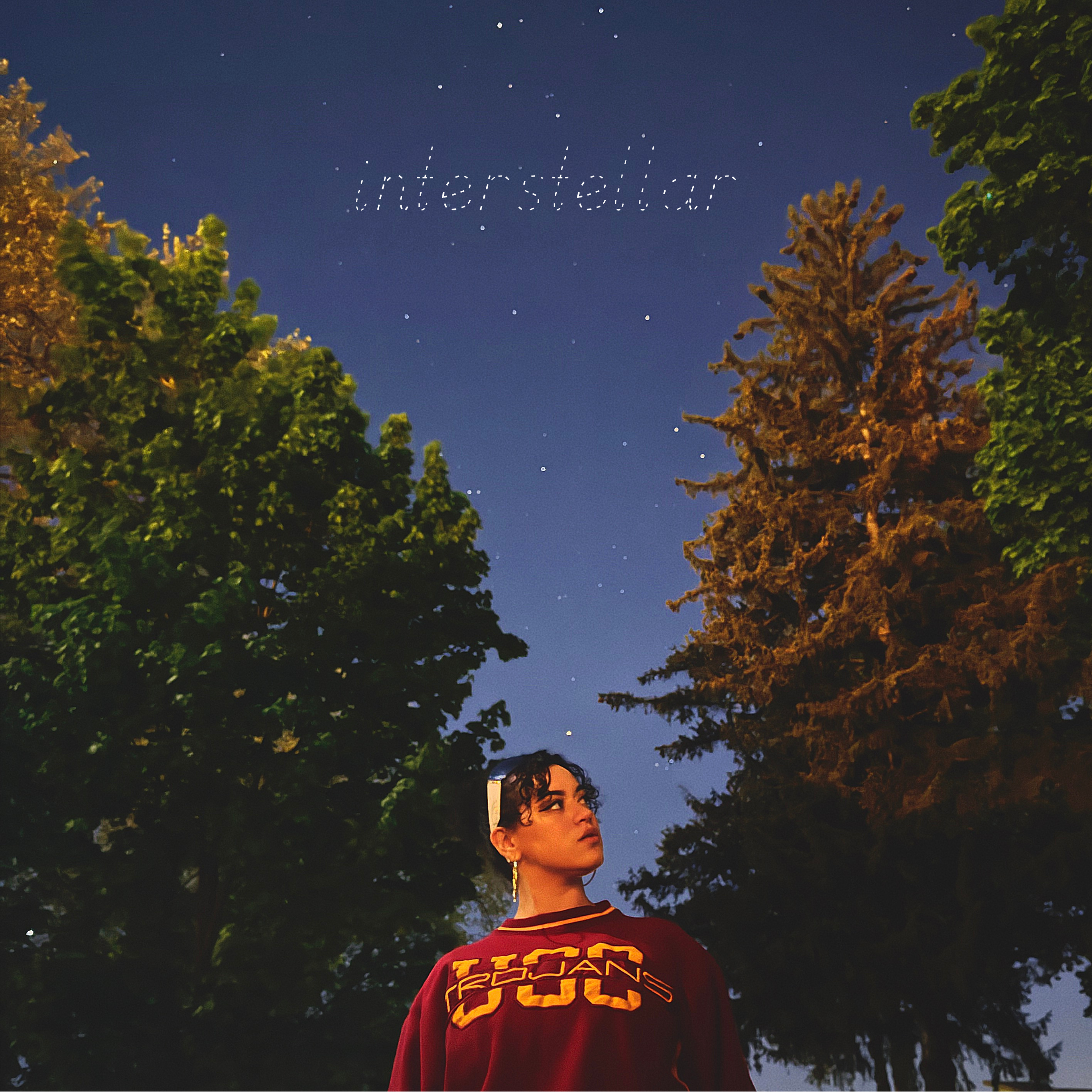 HNY with curly dark hair and earrings stands outdoors at night among trees with a dark blue starry sky overhead. The word 'intersellar' appears in the sky in dotted lines.
