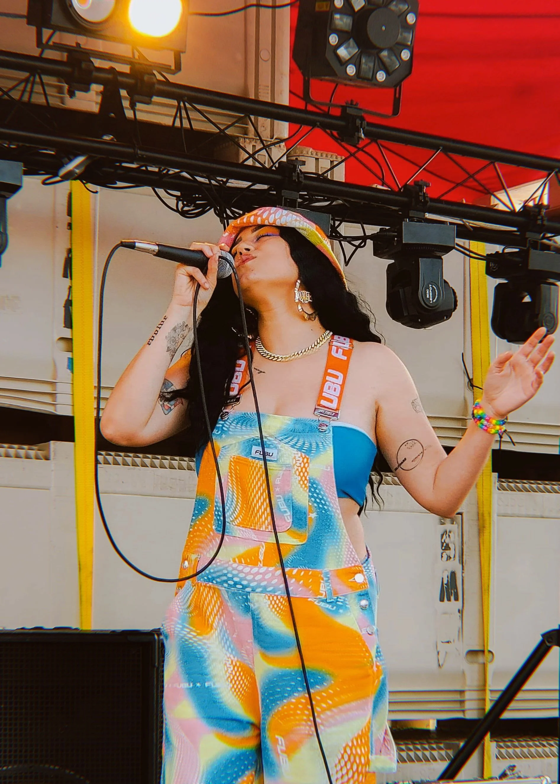 HNY with long dark hair wearing colorful overalls and a bucket hat, singing into a microphone on stage with stage lighting and equipment in the background.