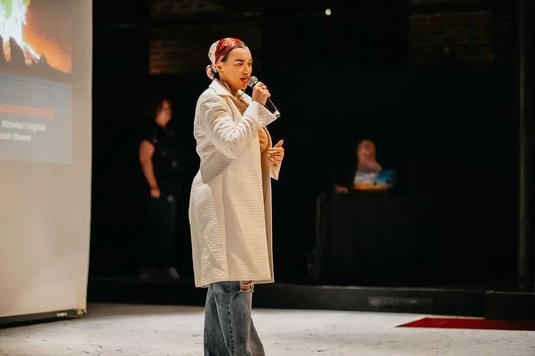HNY woman with a headscarf and beige coat speaking into a microphone on a stage at Gesa Powerhouse Theatre, with a dark background and a person standing in the back.