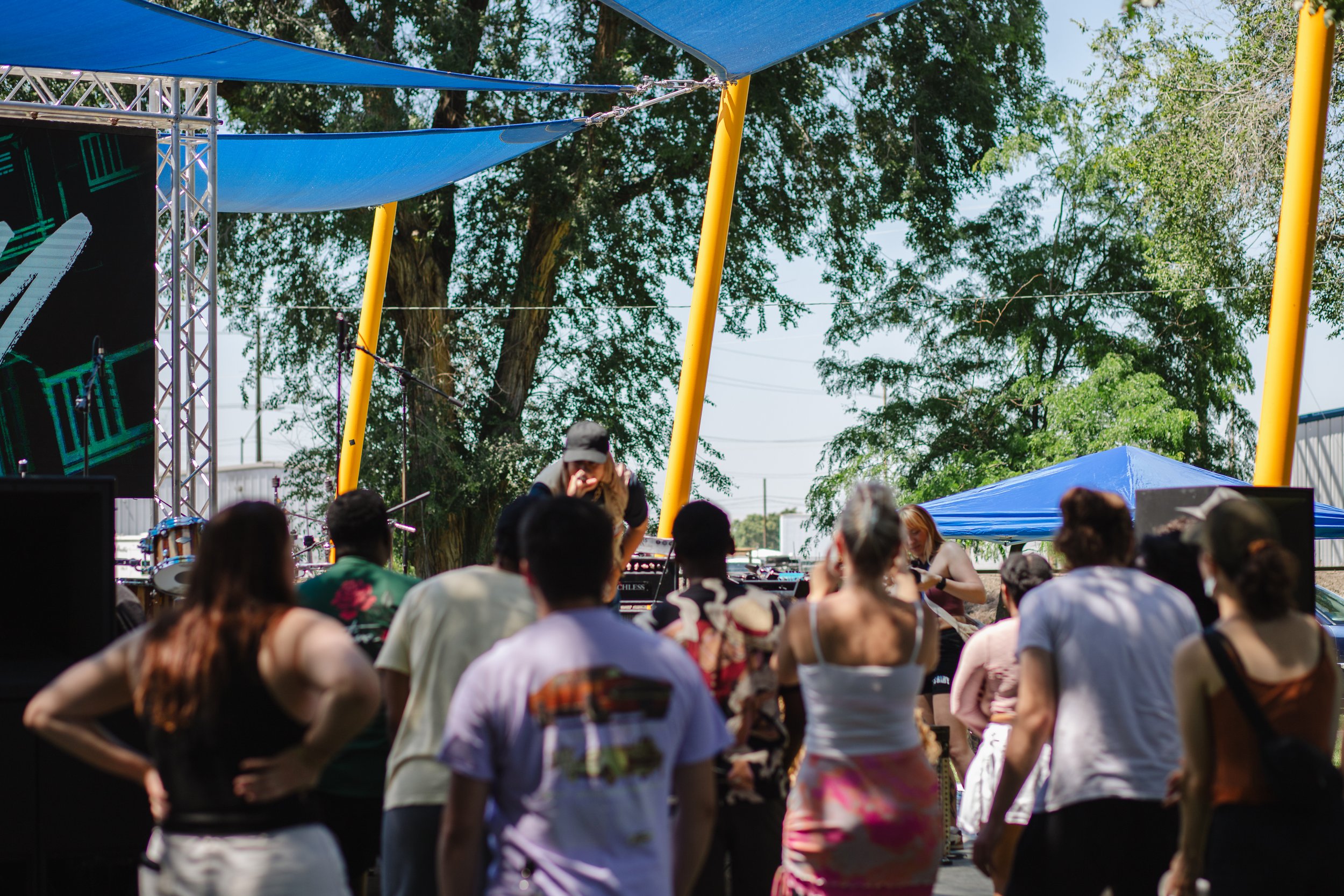 Juneteenth Jubilee Walla Walla with a large crowd, a stage with musical equipment, and HNY wearing a hat, under a canopy with large trees in the background on a sunny day.