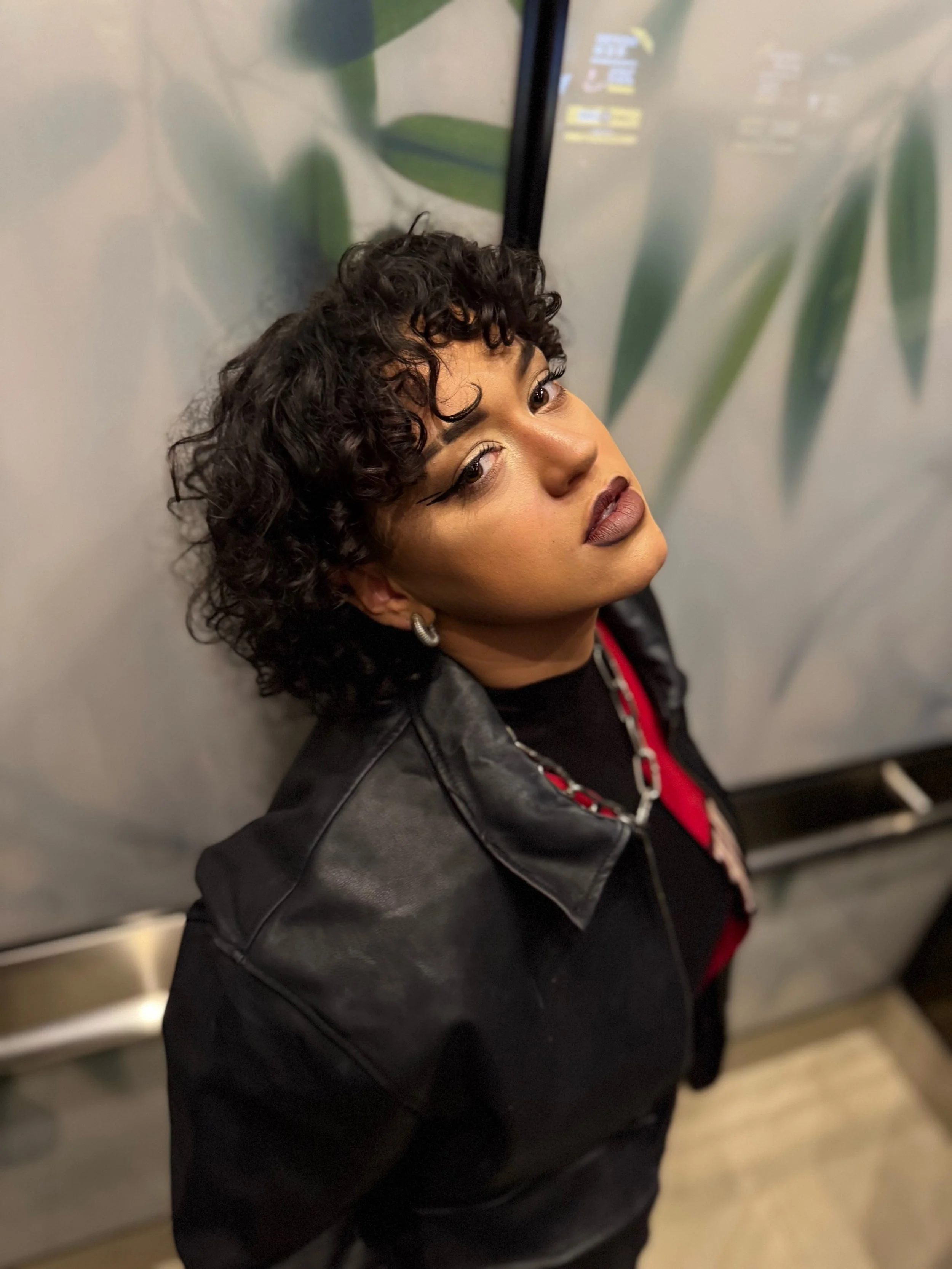 HNY with curly dark hair, makeup, earrings, and a chain necklace leaning against a wall in an elevator.