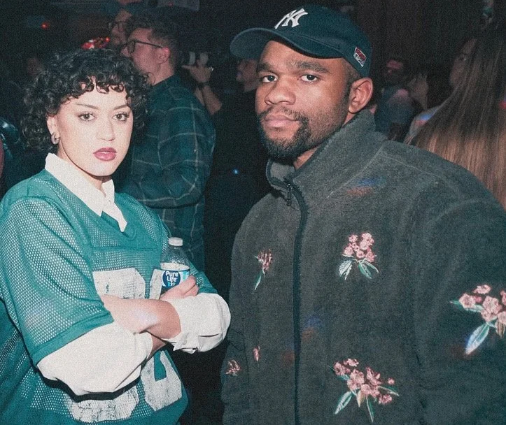 HNY with curly hair wearing a teal sports jersey and holding a water bottle, standing next to Saint Deon wearing a dark floral jacket and a baseball cap, in a dimly lit social setting with other people in the background.