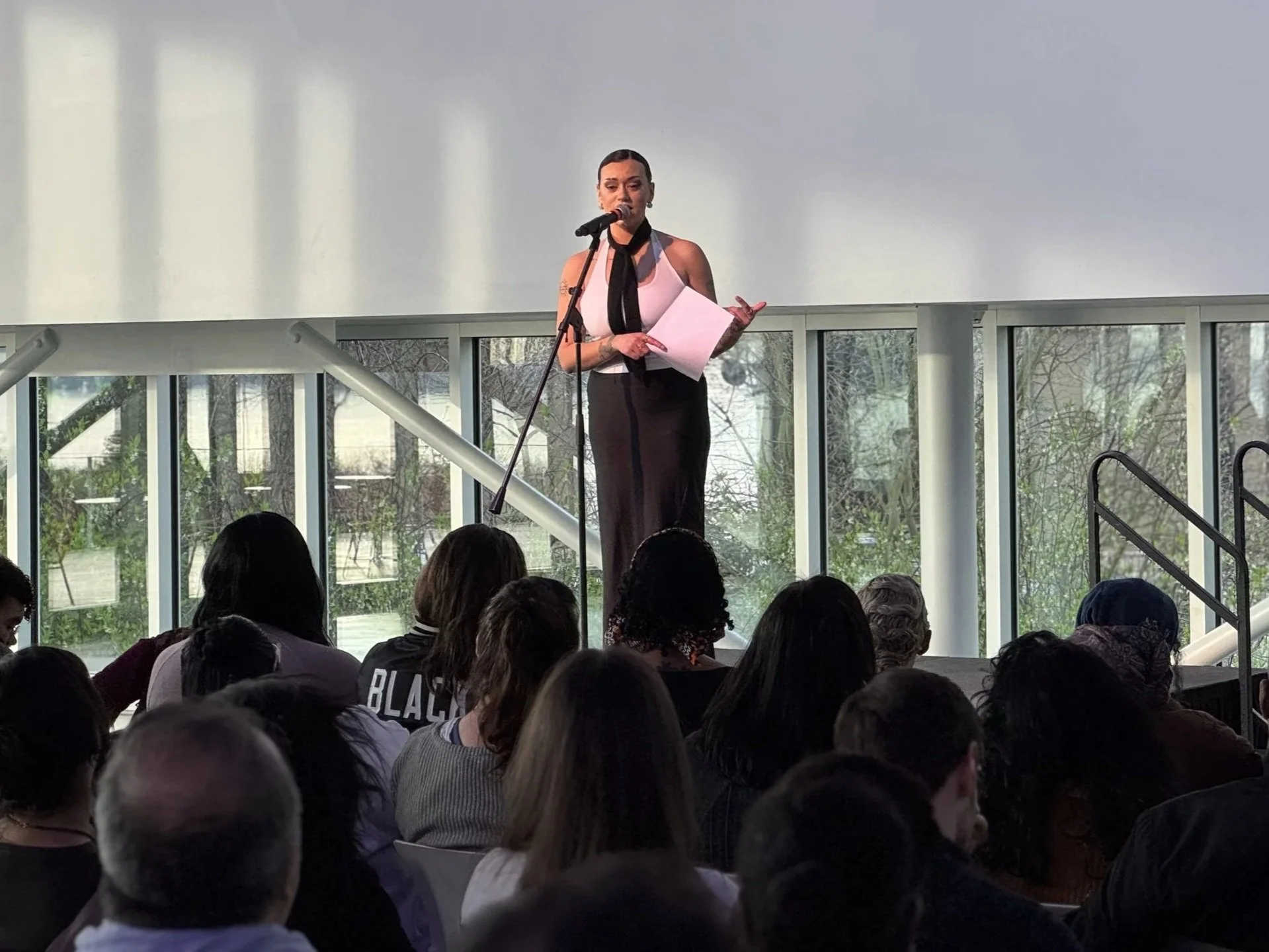 HNY is speaking at a podium at Seattle Art Museum, holding papers, with a crowd seated in front of her and large windows behind her showing greenery.