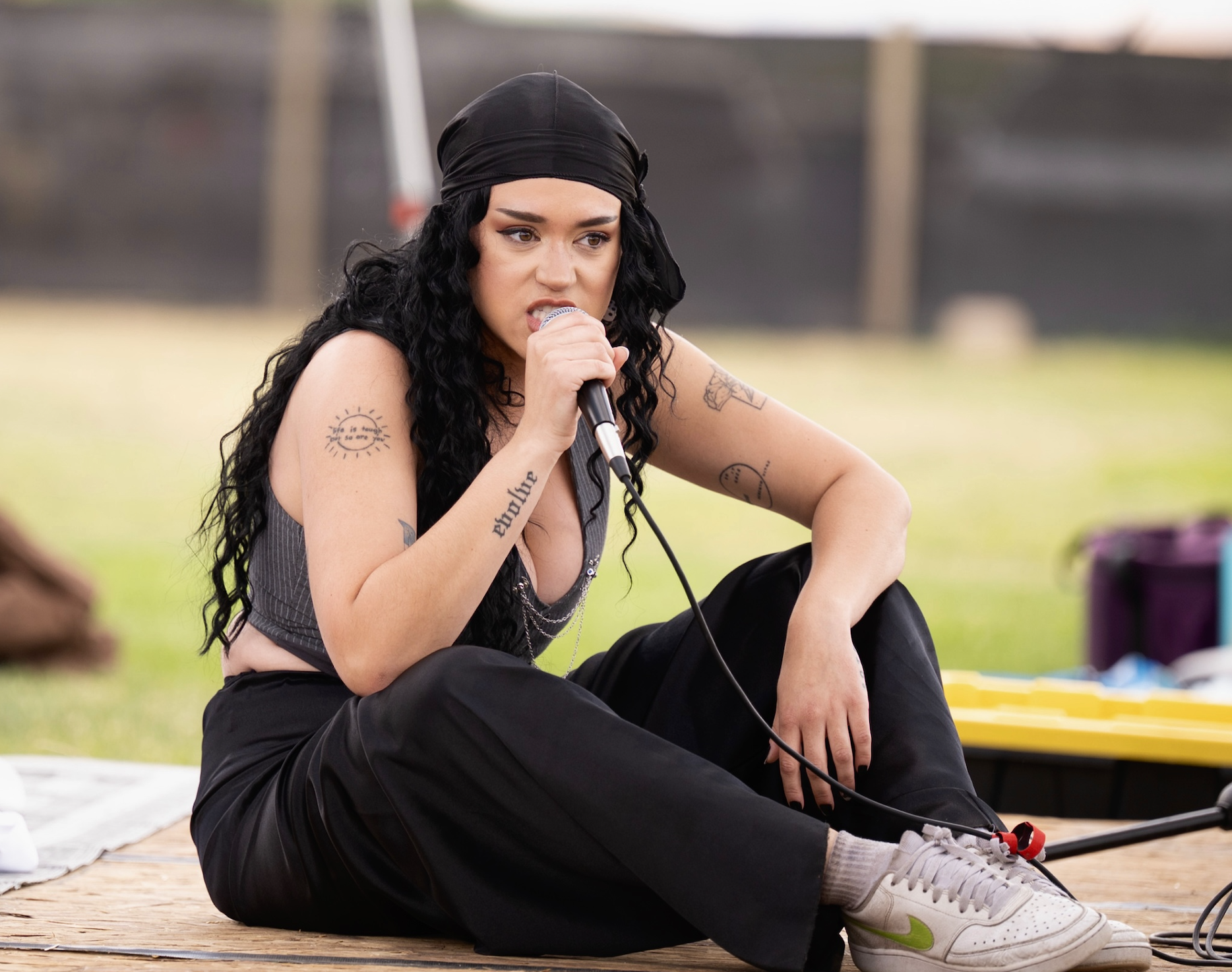 HNY with black curly hair, wearing a black bandana, a gray sleeveless top, black pants, and white sneakers with green accents, sitting on a wooden platform outdoors, holding a microphone and singing or speaking.