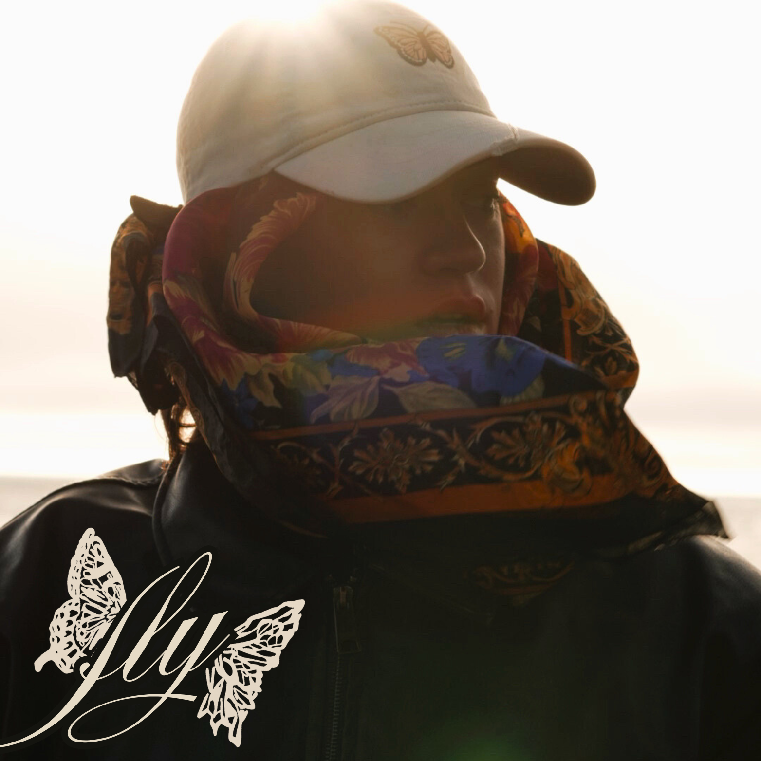 HNY wearing a white baseball cap with a butterfly emblem, a colorful scarf around their neck, and a black jacket with a butterfly and the word 'fly' on it, standing outdoors during sunset.