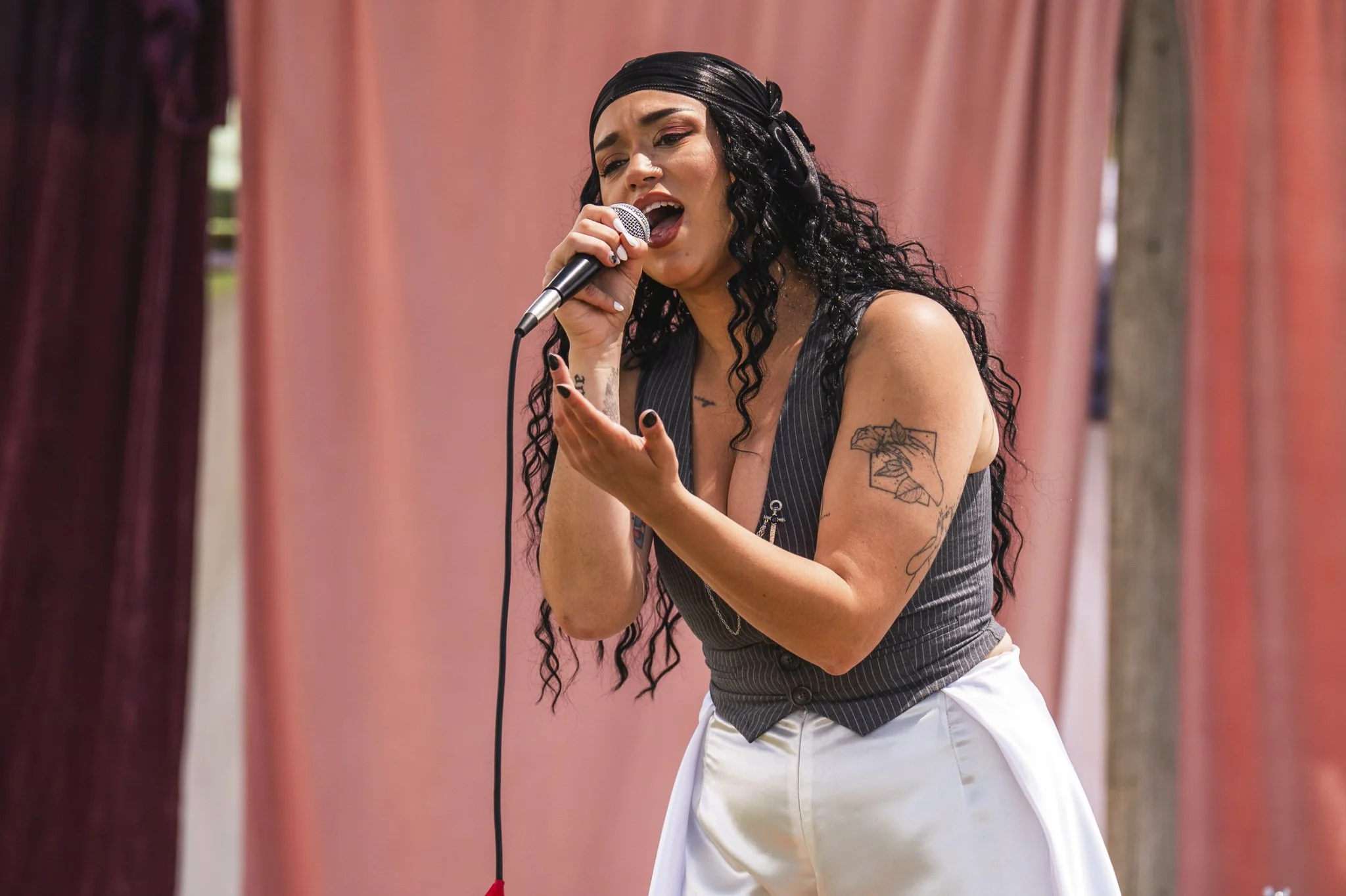 HNY with dark, curly hair sings into a microphone during a performance, wearing a black headband, a sleeveless pinstripe vest, and white pants. She has tattoos on her left arm.