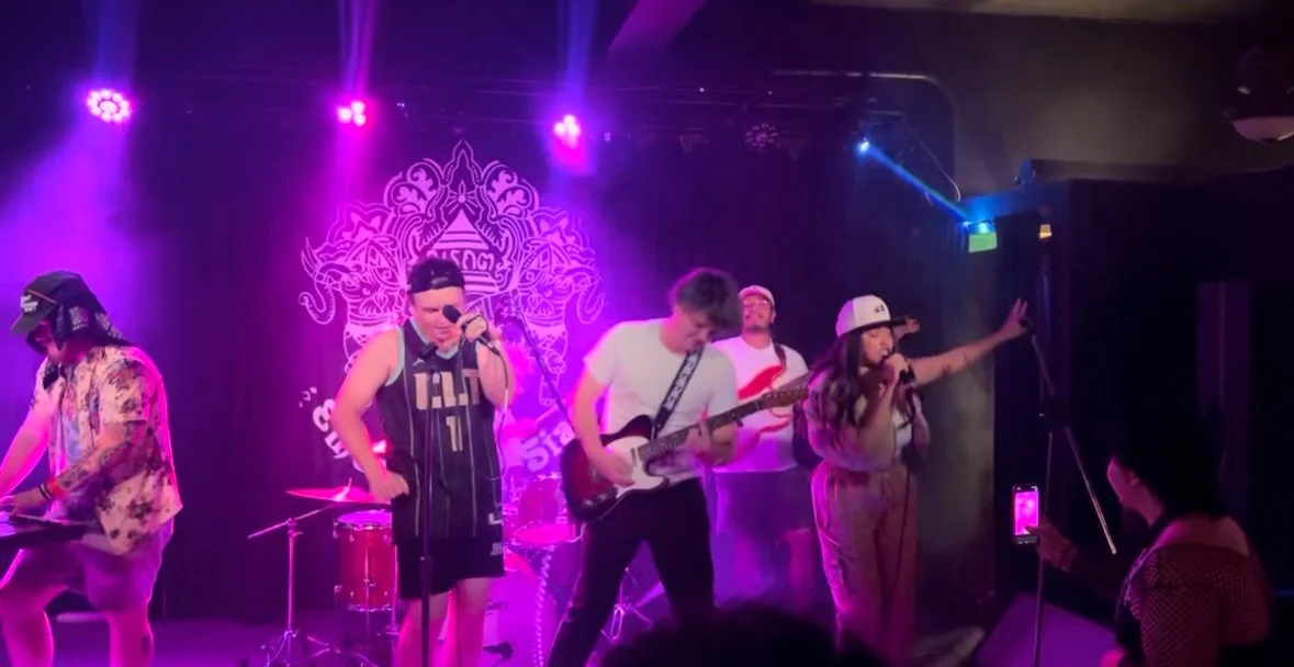 Gravity Well band performing on a stage with purple and pink lighting, members playing guitar, keyboard, and singing, with a decorative backdrop.